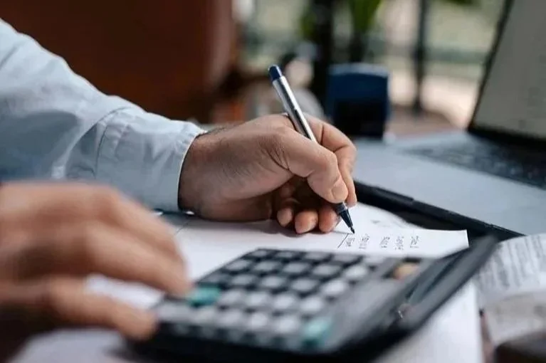 man sitting at desk next to large window writing down numbers while typing on calculator