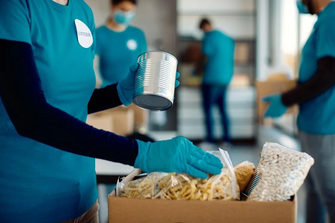 Nonprofit volunteer wearing gloves and a blue t-shirt packs up groceries in a cardboard box while other volunteers work in the background.