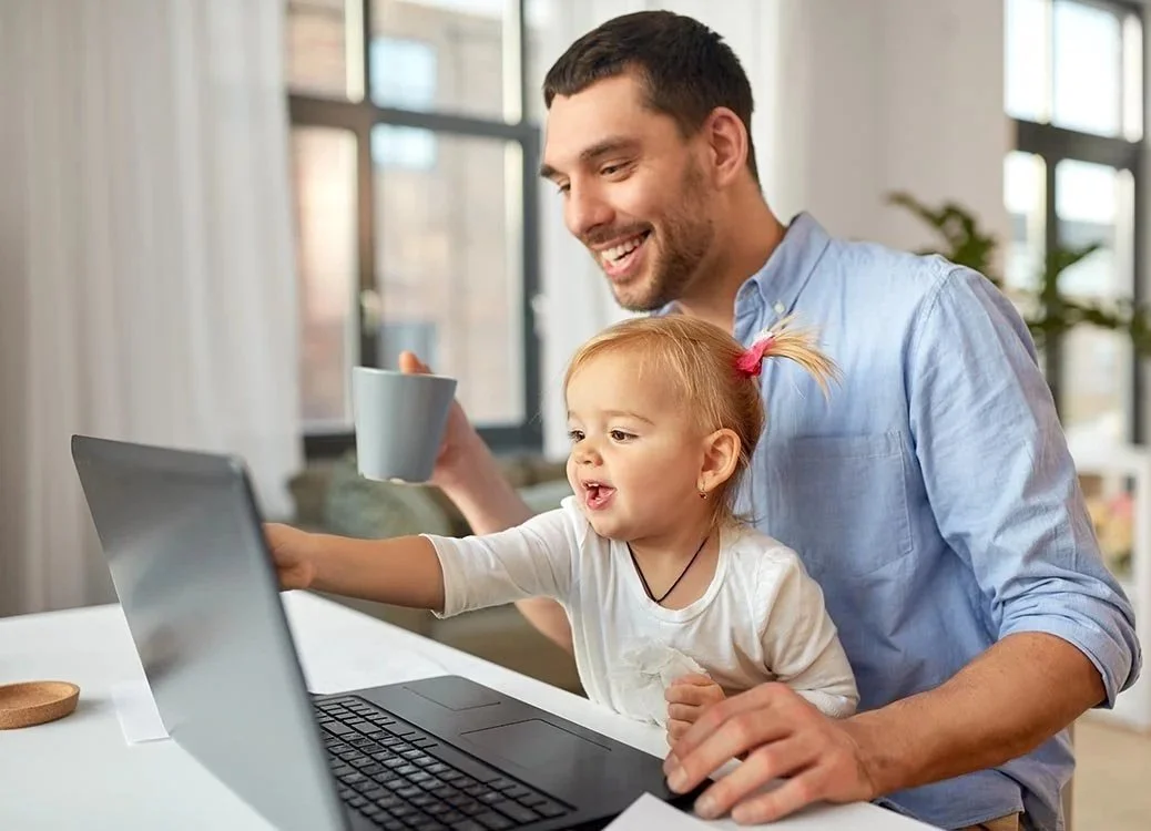 Dad smiling while holding a coffee cup and working on laptop with young daughter sitting in his lap pointing on his computer screen.