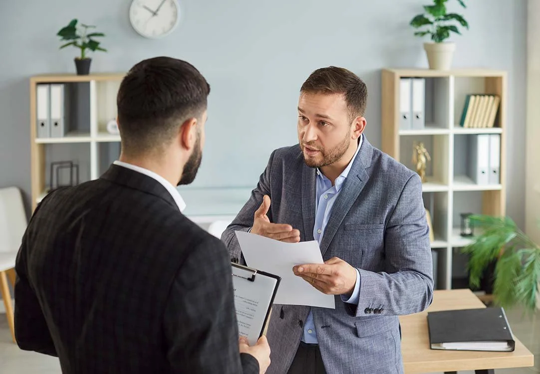 Upset employee holding a paper and gesturing while talking to a manager holding a clipboard.