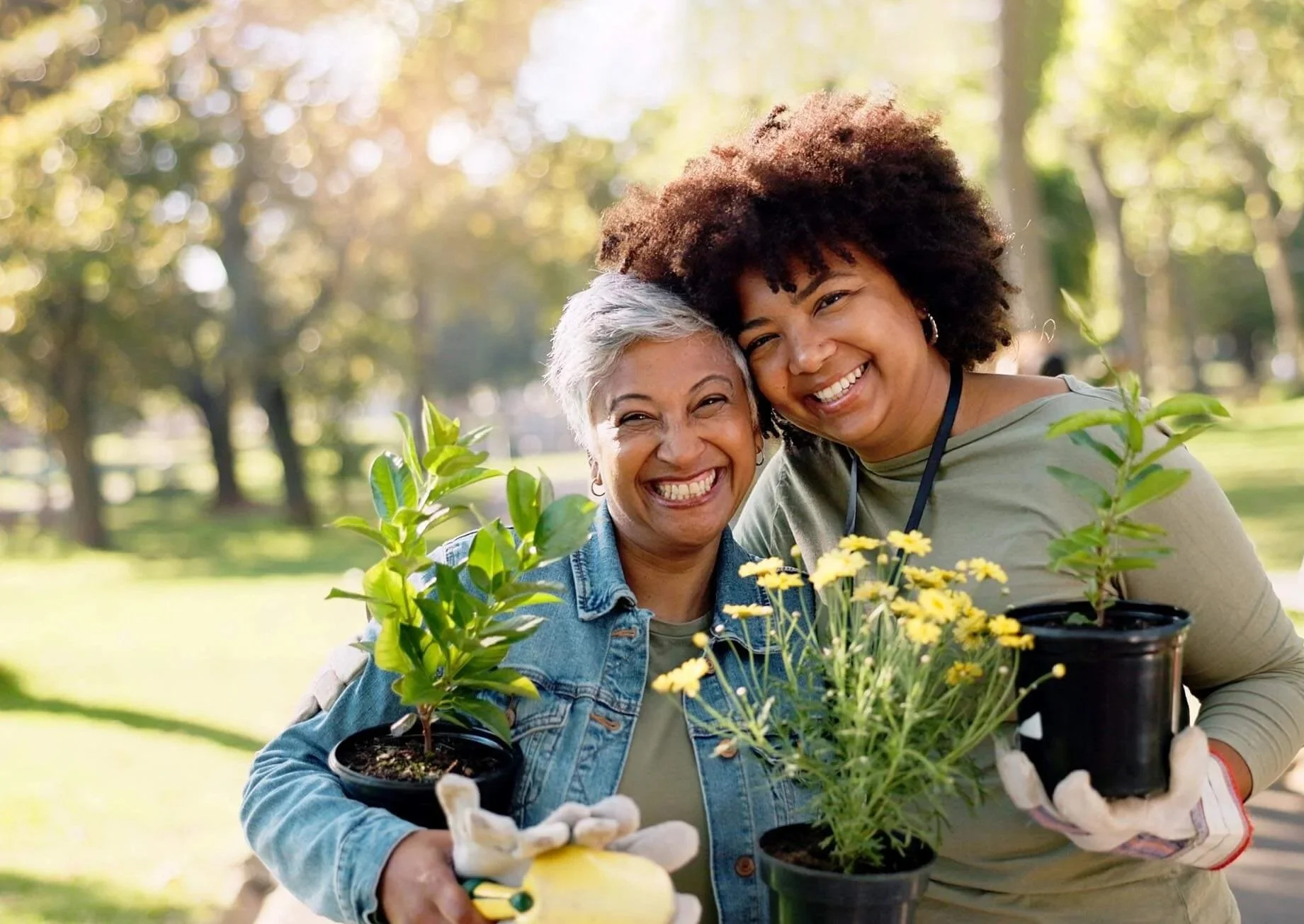 Two smiling women in a park hugging from the side while holding plants in pots during community event.