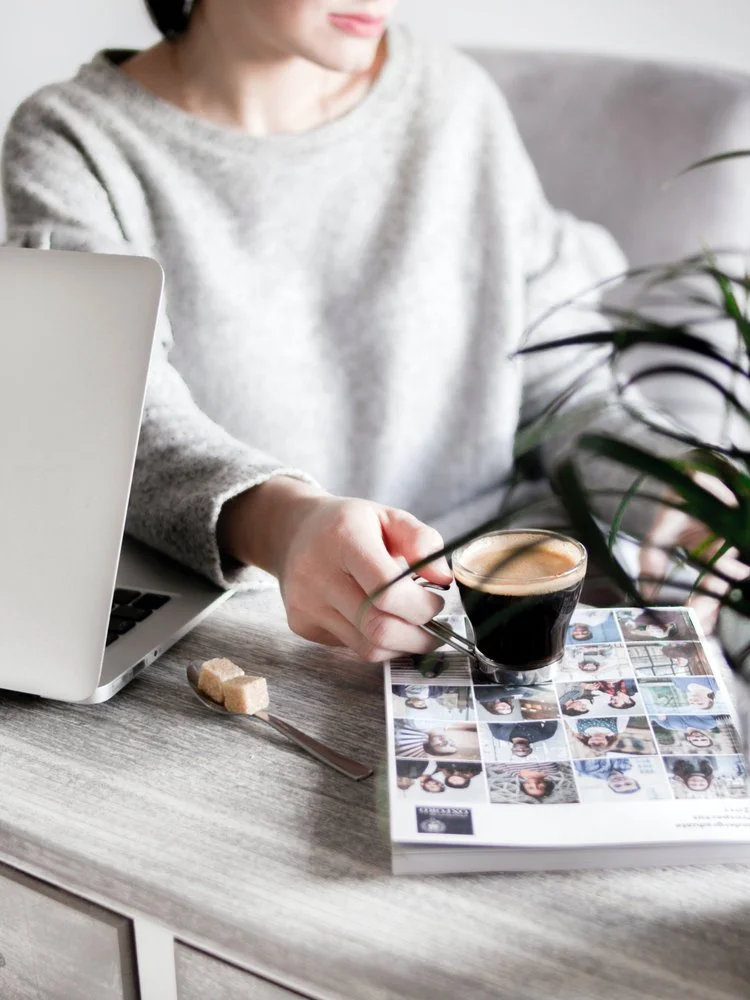Person in a gray sweater sitting at a desk with a laptop, a spoon with sugar, a small glass of coffee, and a photo collage magazine, with a green plant partially visible in the foreground.