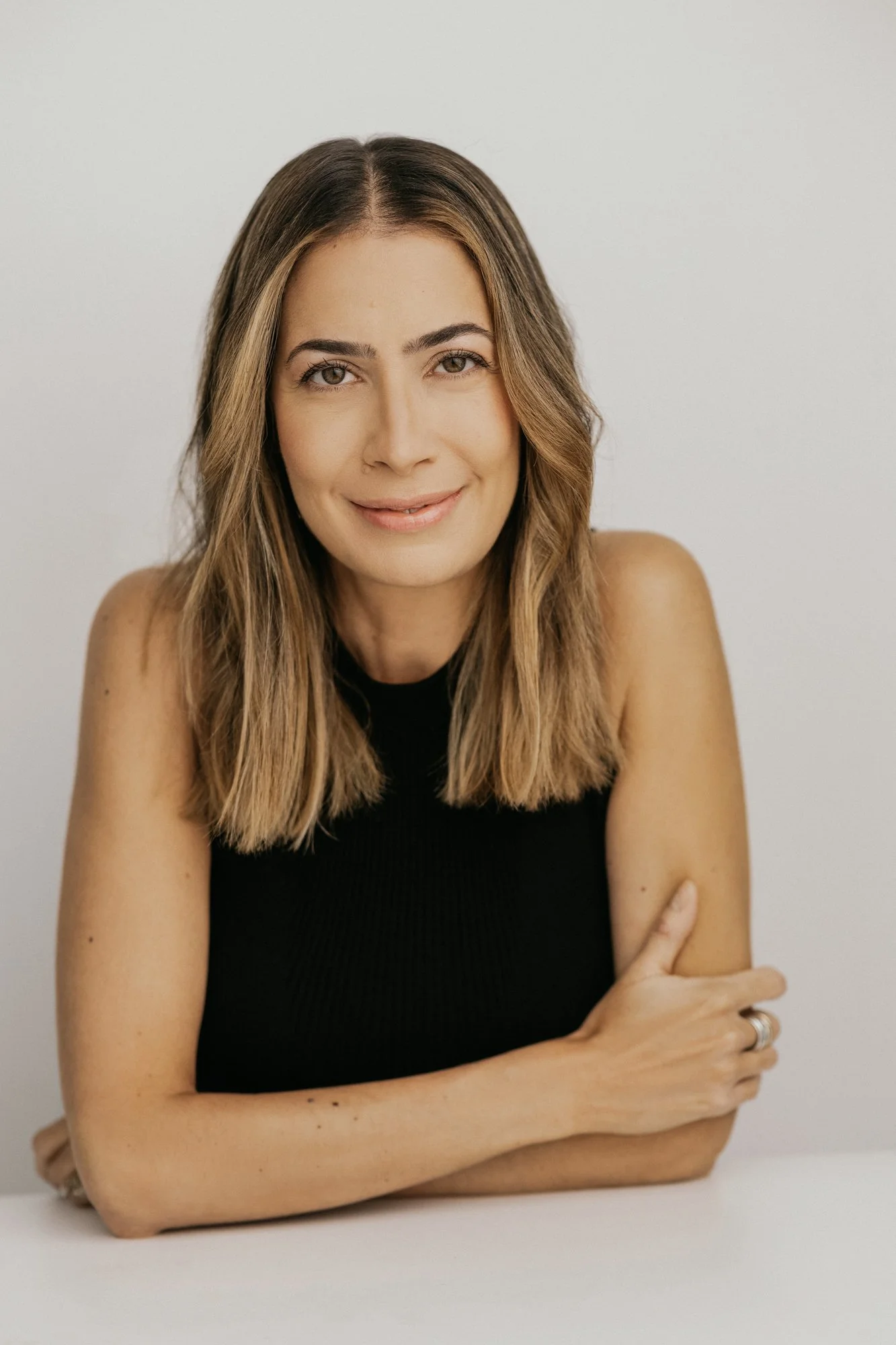 A woman with shoulder-length brown hair, wearing a sleeveless black top, smiling and looking at the camera against a plain light-colored background.