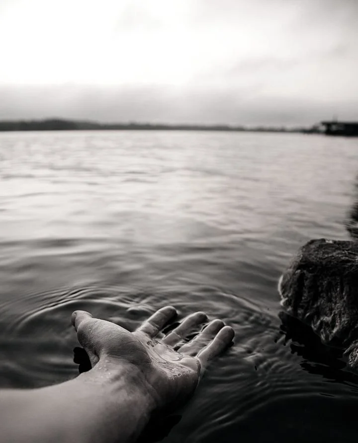 A black and white photo of a hand reaching into a body of water near a rock, with the water and sky in the background.