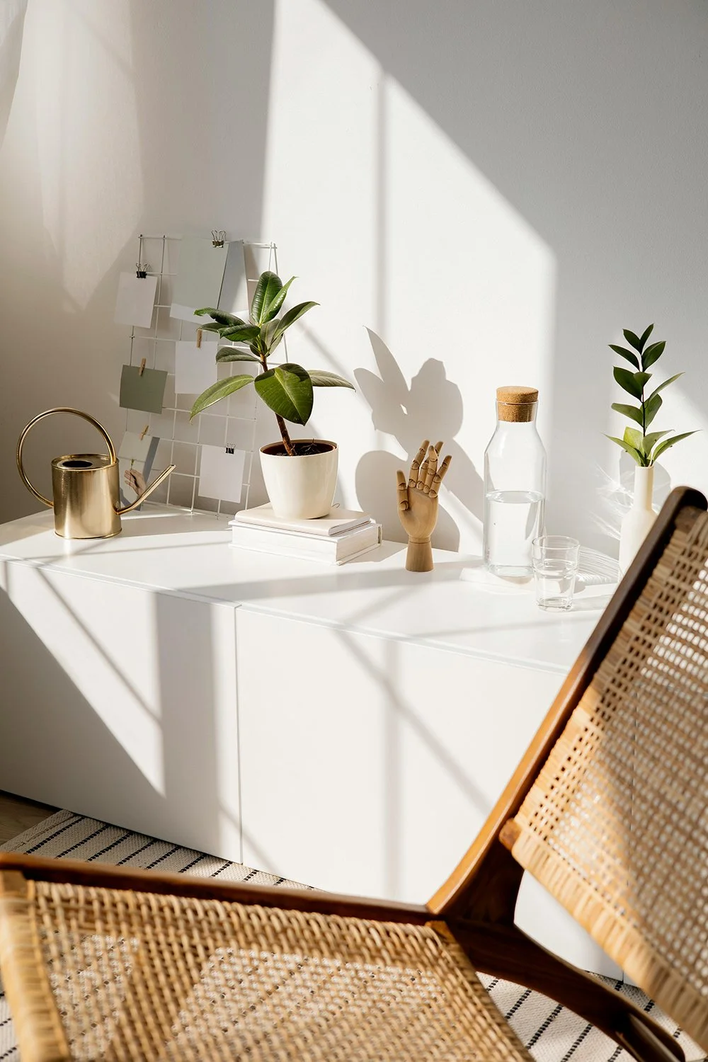 Sunlit corner of a modern interior space with a white cabinet, potted plant, glass water jug, glass, decorative mannequin hand, and a rattan chair.