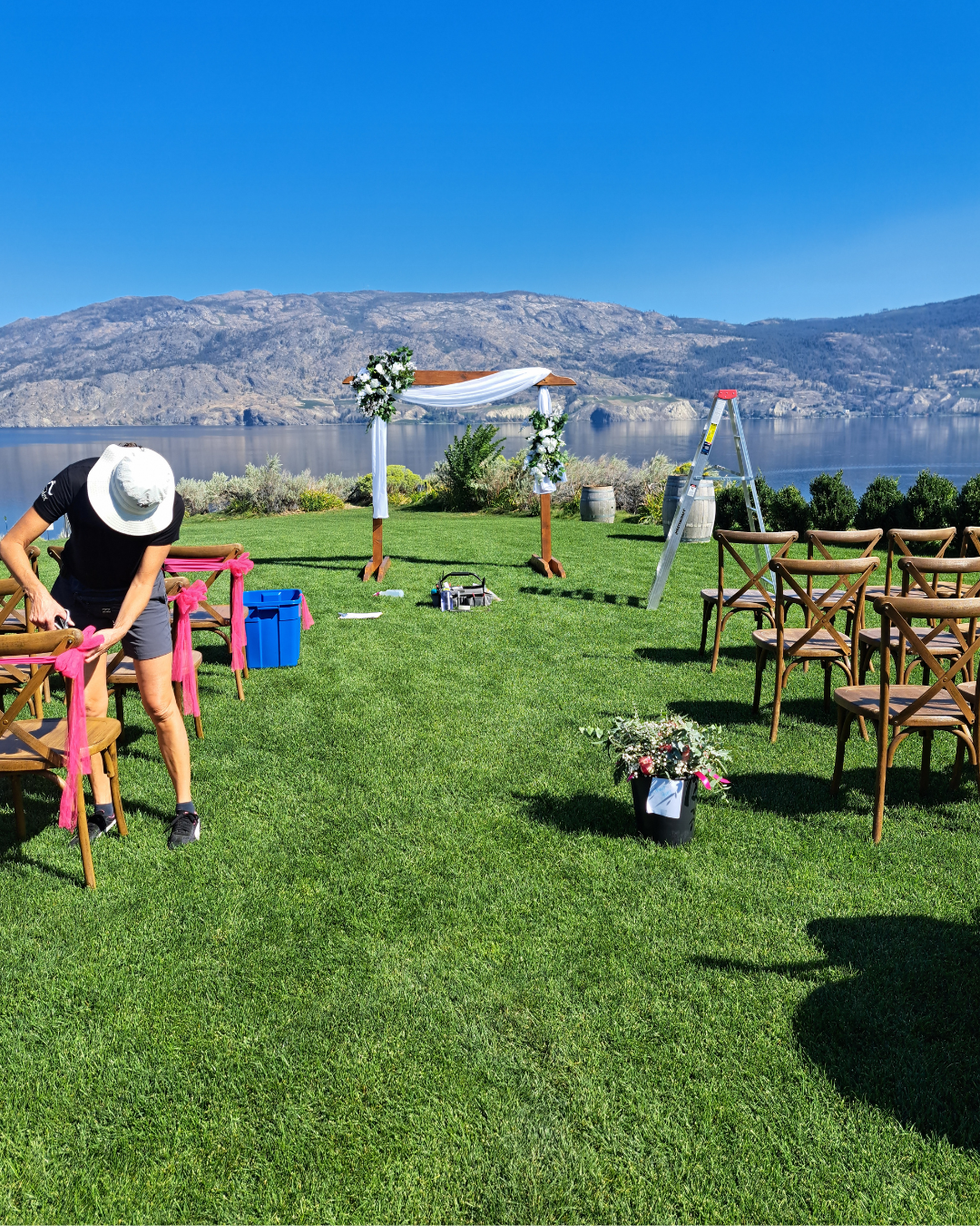 Setup for an outdoor wedding ceremony by a lake with chairs, a decorated arch, and a person working on pink ribbons.