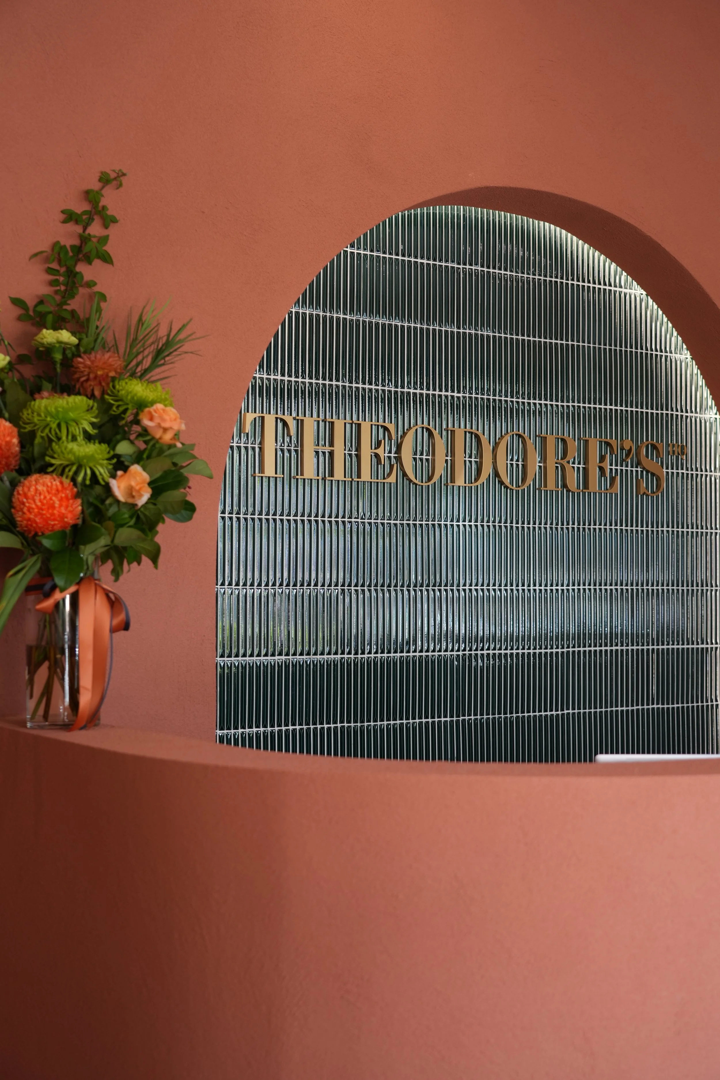 Close-up of a sign reading 'Theodore's' on a metallic background, with a flower arrangement on a pink wall to the side.