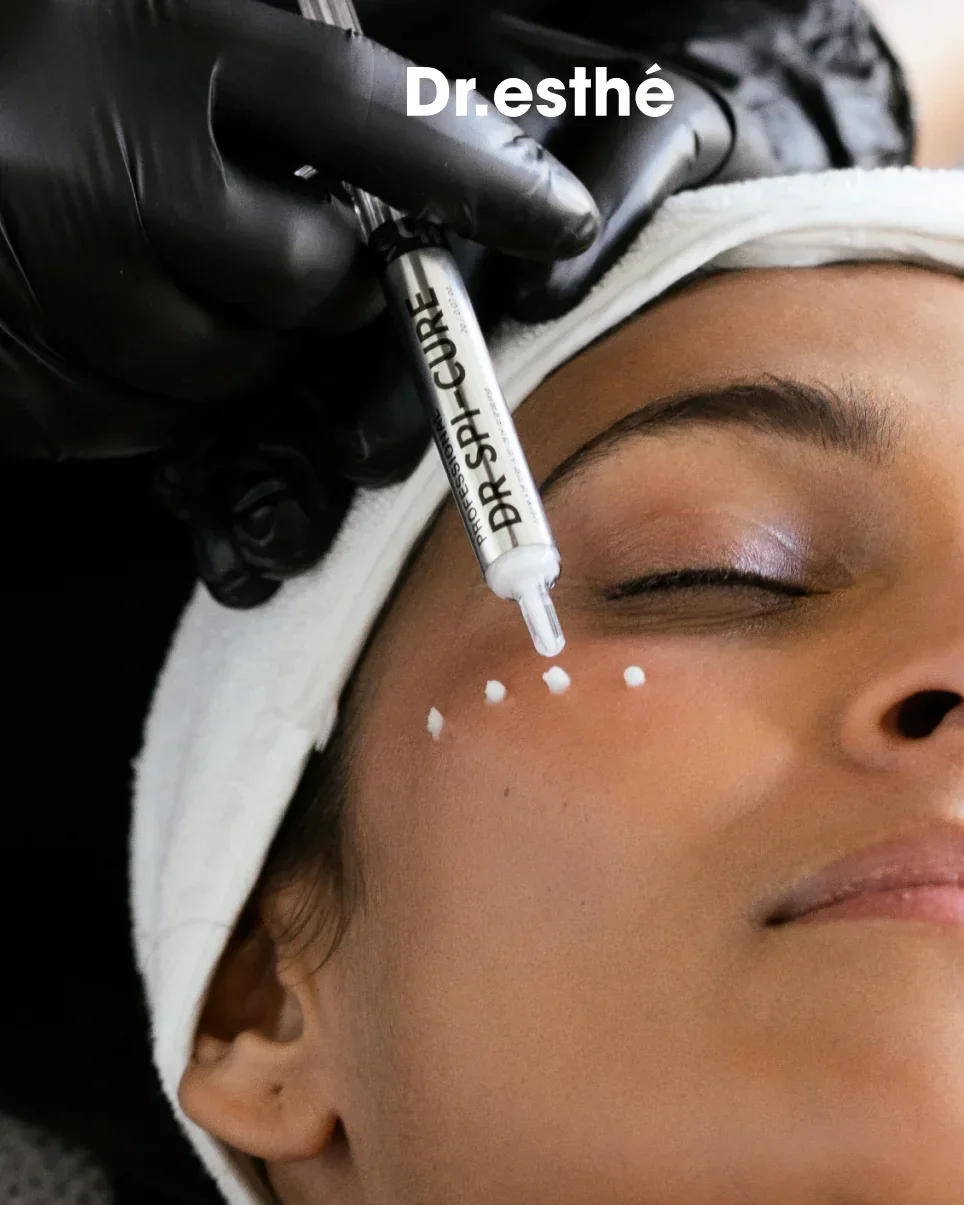 A person getting a facial treatment with small white dots applied near their eye, while a cosmetic professional applies a white serum labeled 'DR. SPARCURE' with a pipette. The person is lying down with their eyes closed, wearing a white headband.
