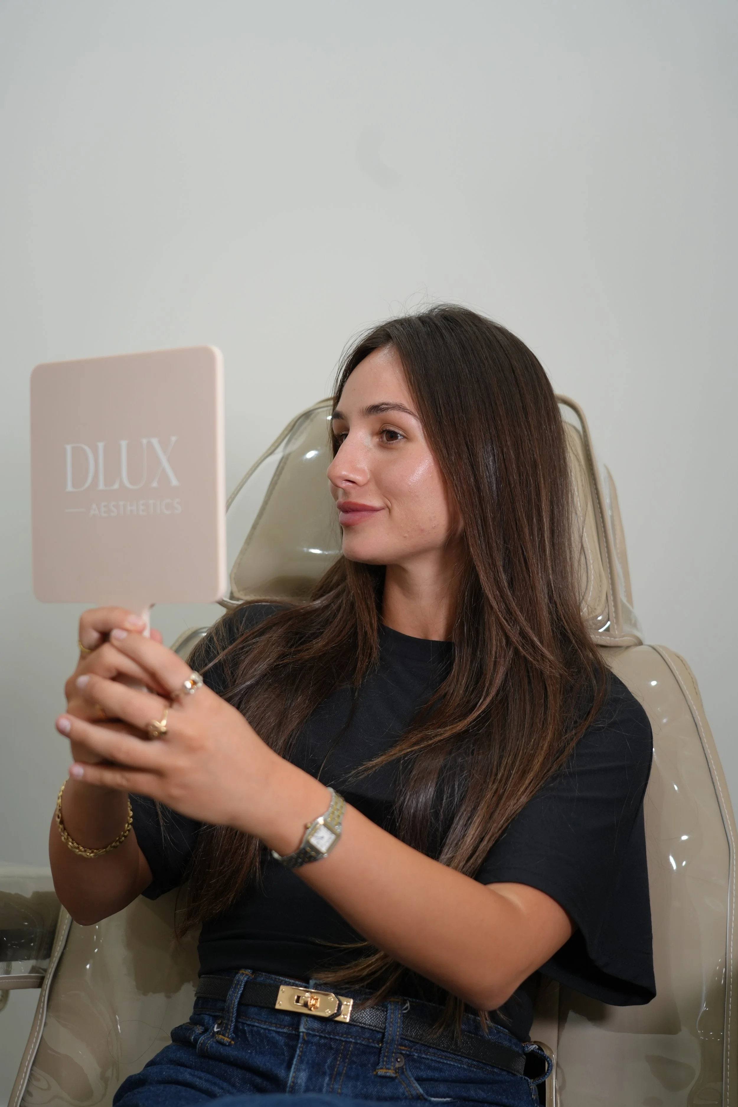 A young woman with long brown hair is sitting on a beige chair, looking at herself in a pink mirror with the words "DLUX AESTHETICS" written on it. She is wearing a black shirt, blue jeans with a black and gold belt, and gold jewelry. She appears to be doing a skincare or beauty routine and is in a room with plain light-colored walls.