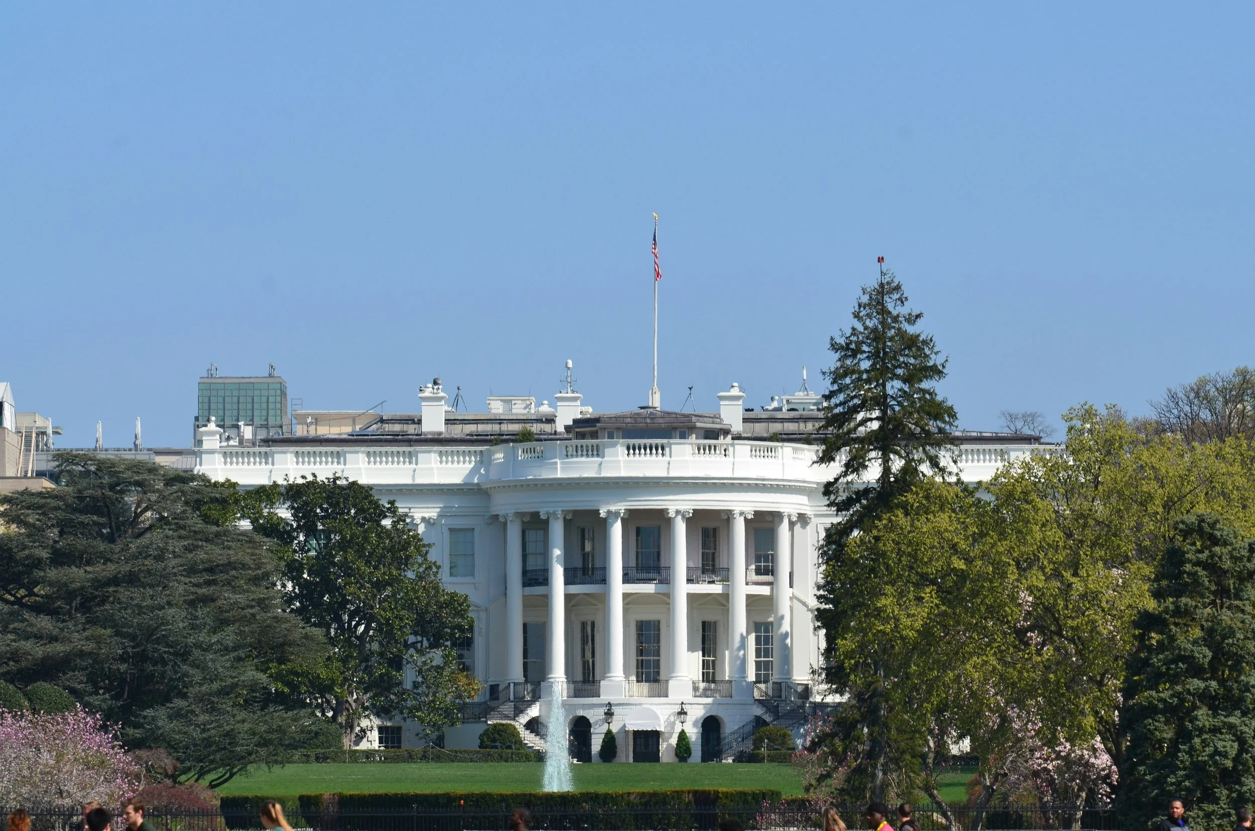 The White House, a large white neoclassical building with tall columns, is surrounded by trees and greenery. An American flag is flying on a tall flagpole on the roof, and there is a fountain in front of the building. People are visible in the foreground.