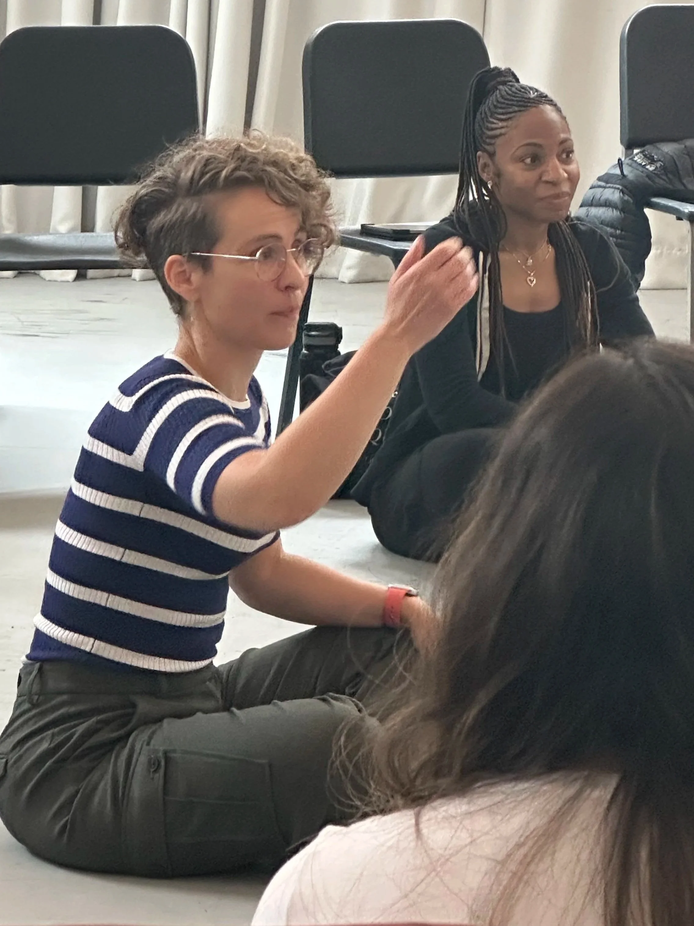 A young woman with short curly hair, glasses, and a striped shirt sitting on the floor, raising her hand while speaking. An older woman with long braids, wearing a black jacket, listens attentively.