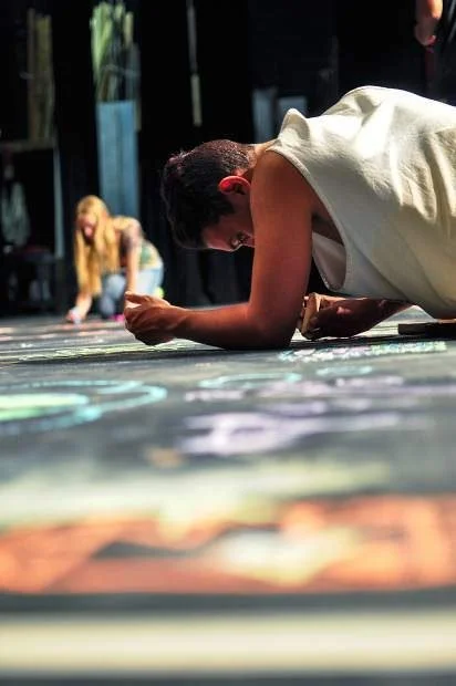 Person lying on the floor drawing with chalk, with another person in the background also drawing.