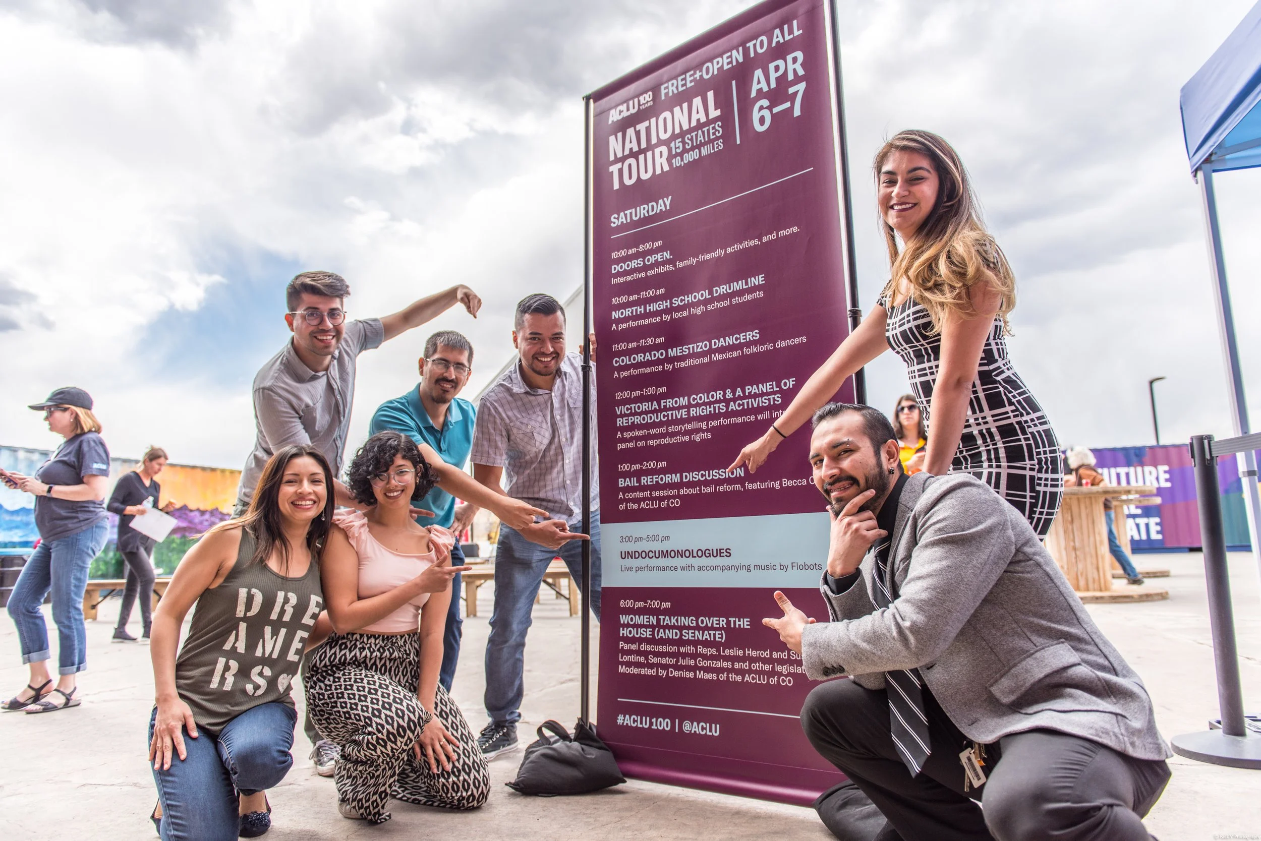 Group of people smiling and pointing at a large event schedule sign at an outdoor festival on a cloudy day.