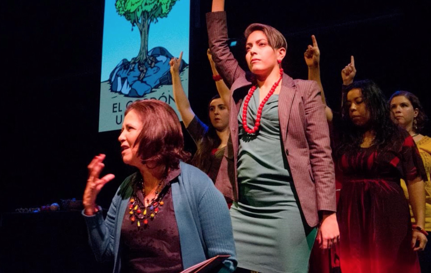 A group of women on stage, some raising their fists. One woman in front with short hair and a grey blazer is speaking, while another woman with short hair, red necklace, and green dress is standing beside her with her fist raised. A large screen behind them displays a drawing of a tree and the words "EL" and "U".