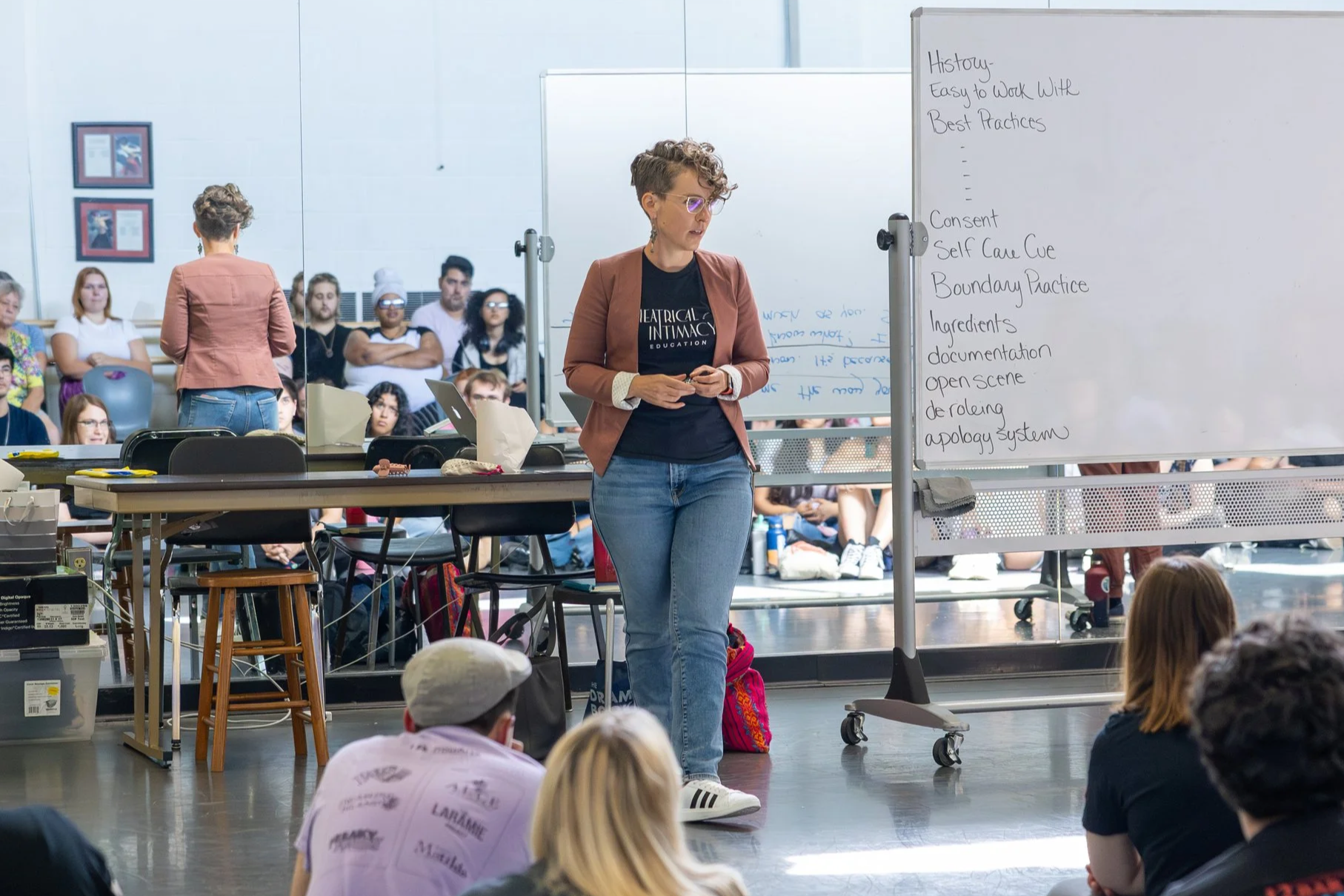 A woman standing at the front of a group of seated people, giving a presentation or lecture. She is wearing glasses, a brown blazer, a black T-shirt, and blue jeans. Behind her, there is a whiteboard with notes about history and best practices. The audience appears attentive and engaged.