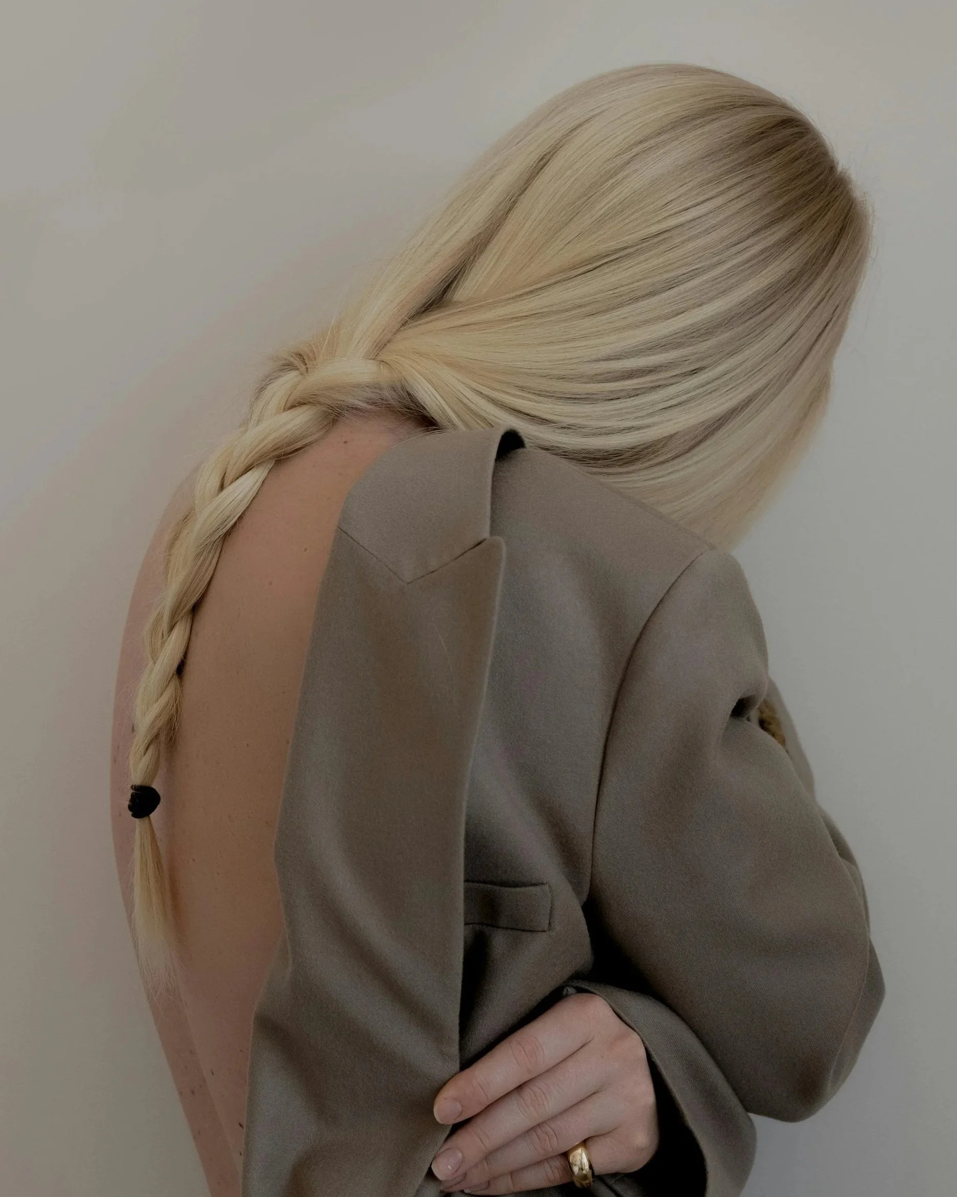 A woman with long blonde hair in a braid, wearing a taupe blazer, partially revealing her bare back.