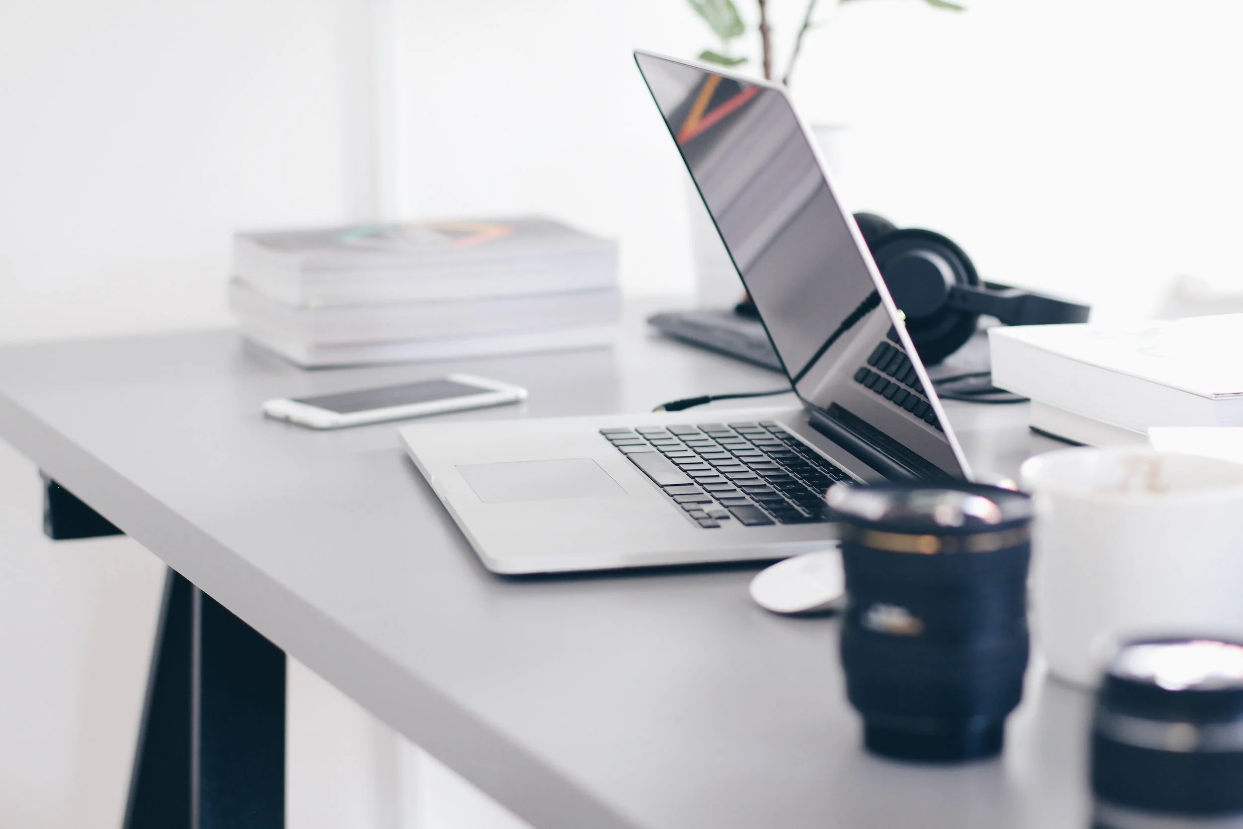 A modern white desk with an open silver laptop, smartphone, black camera, headphones, and stacked notebooks.
