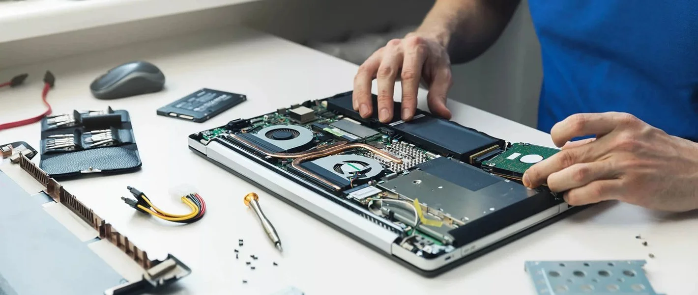 Person repairing a laptop with its cover removed, exposing internal components on a white work surface with tools and parts nearby.
