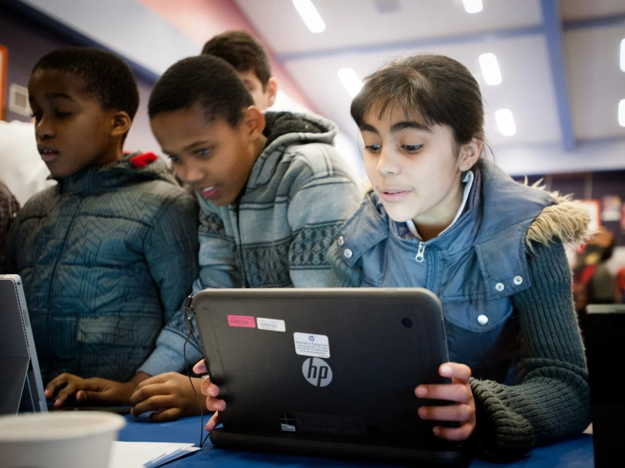 Group of children working together on laptops in a classroom or library setting.