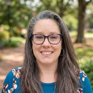 A woman with long dark brown hair speckled with gray,  a dark teal shirt, and glasses smiling outdoors in a park with trees in the background.