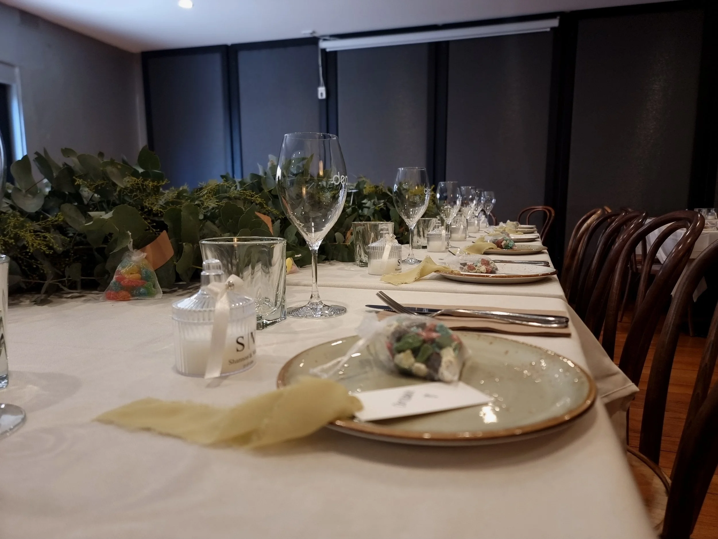A long dining table set for a meal, decorated with greenery and candles, with empty plates, glasses, and utensils, and candy bags on the table.