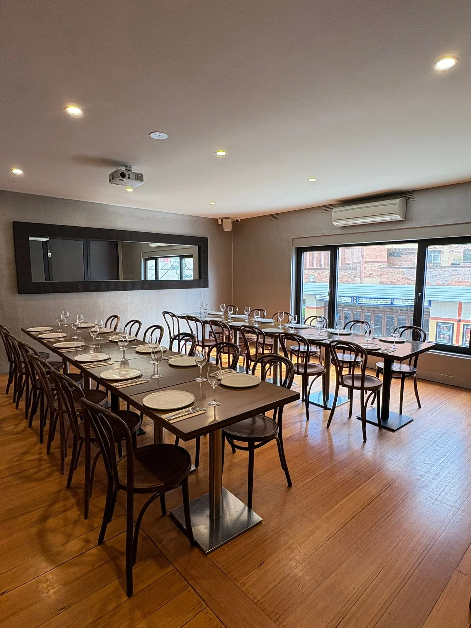 Dining room with a long table set with white plates, wine glasses, and silverware, surrounded by black chairs, large window with city view, wall mirror, ceiling lights, and an air conditioning unit.