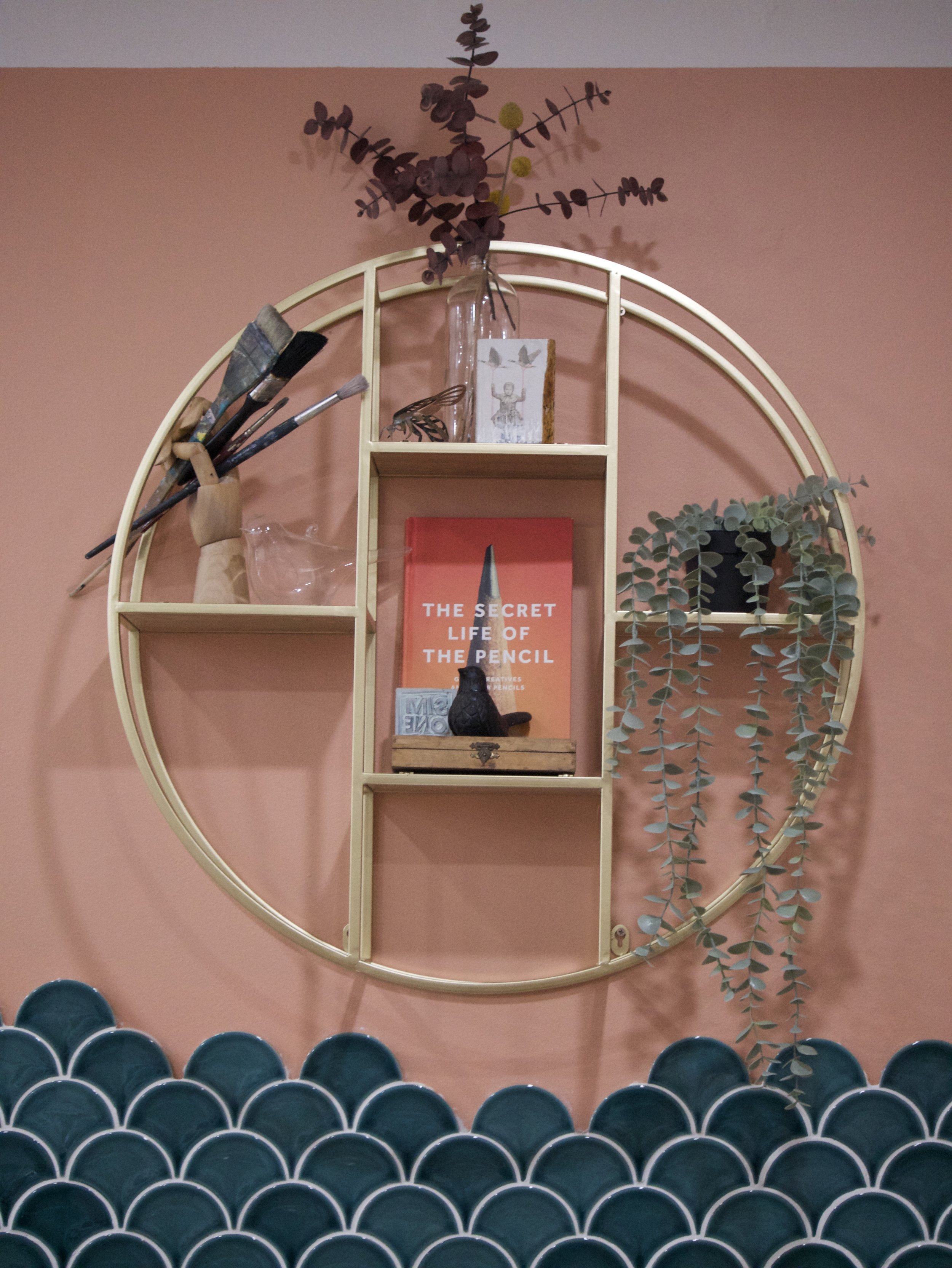 Circular wall shelf with decor items including a red book, paintbrushes, dried eucalyptus, and a small figurine, set against a coral wall with teal scalloped tiles.