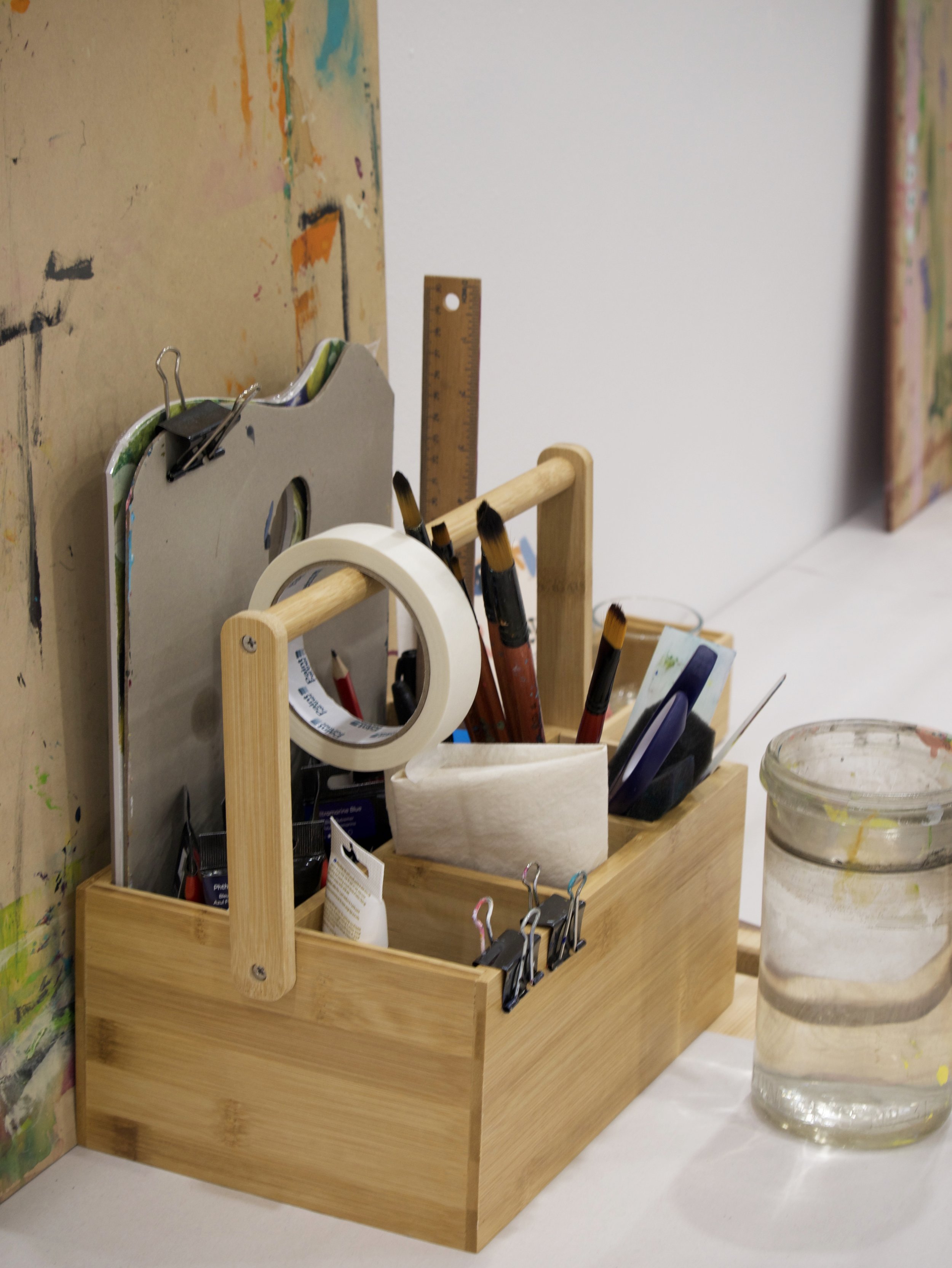 An artist's workspace featuring a wooden organizer holding paintbrushes, masking tape, a ruler, and binder clips. A glass jar filled with water is nearby, and a paint-stained board is in the background.