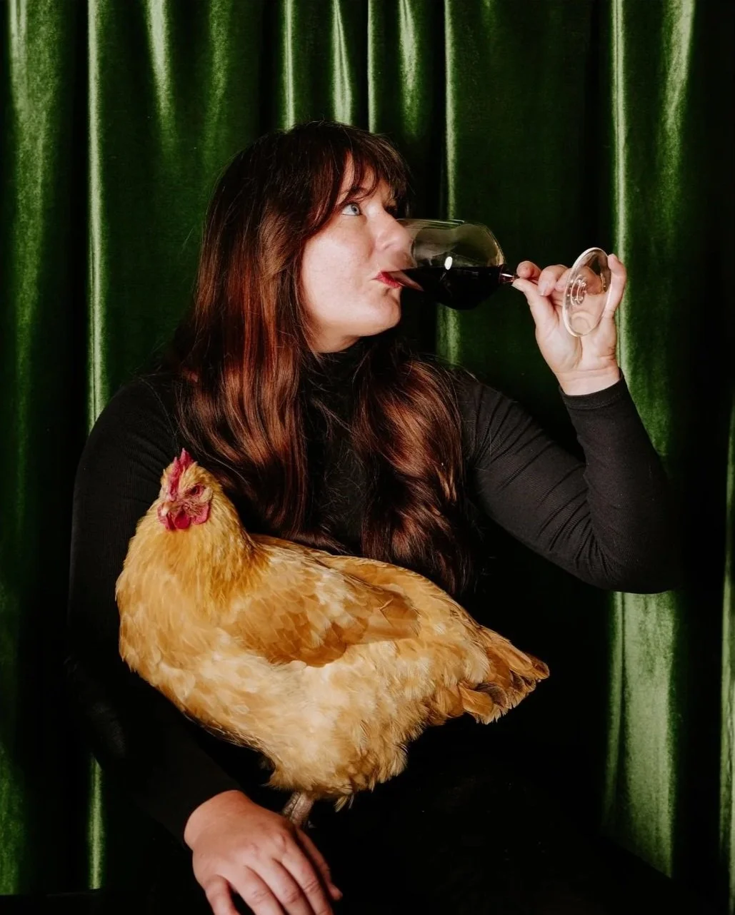 Lifestyle portrait of Heather Daenitz drinking red wine while holding a chicken.