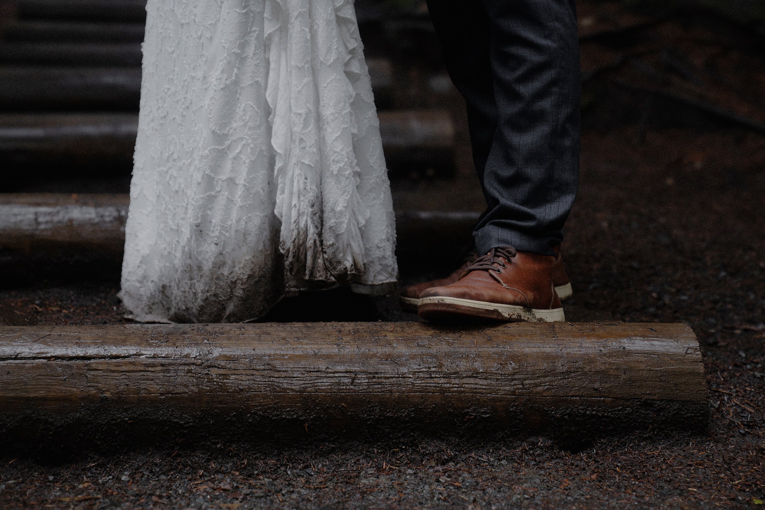 Bride in wedding dress standing on wooden log next to man in suit and brown leather shoes.