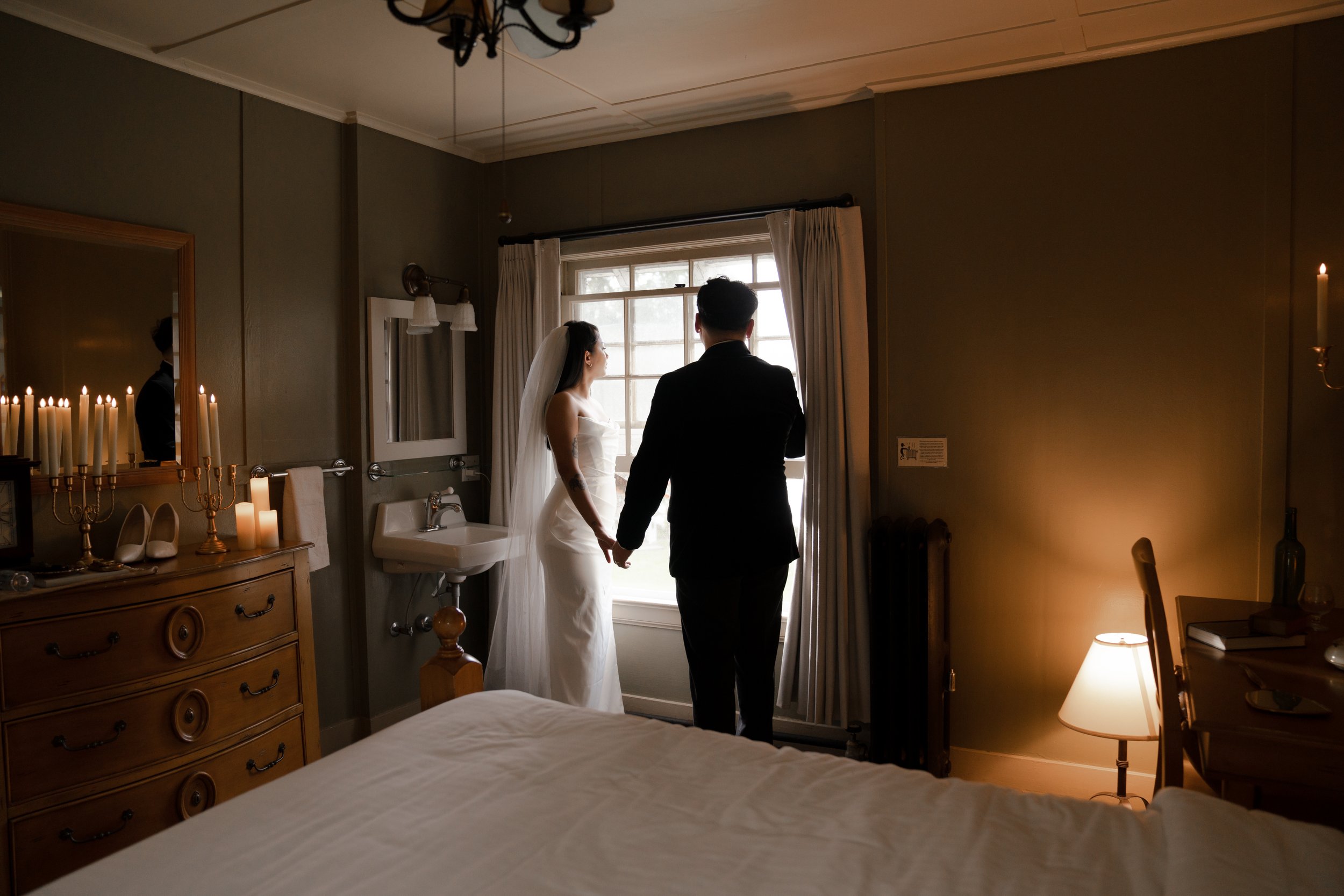 Bride and groom holding hands, standing near a window in a warmly lit room with candles, dresser, and a bed.