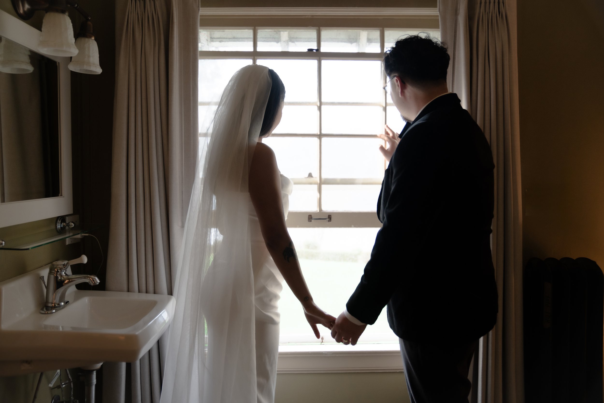 A bride and groom holding hands facing a window, with the bride wearing a white dress and veil, and the groom in a dark suit.