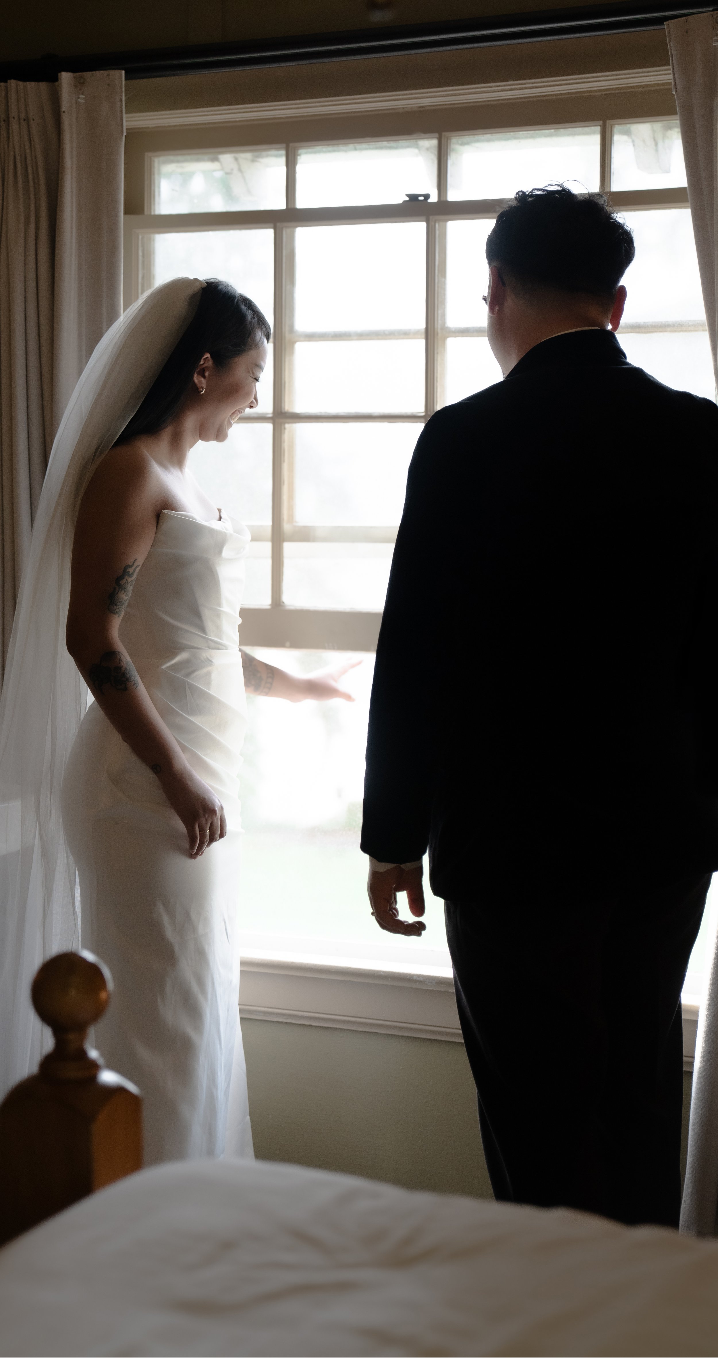 Bride in white dress and veil stands with a man in a dark suit, looking out a window with curtains.