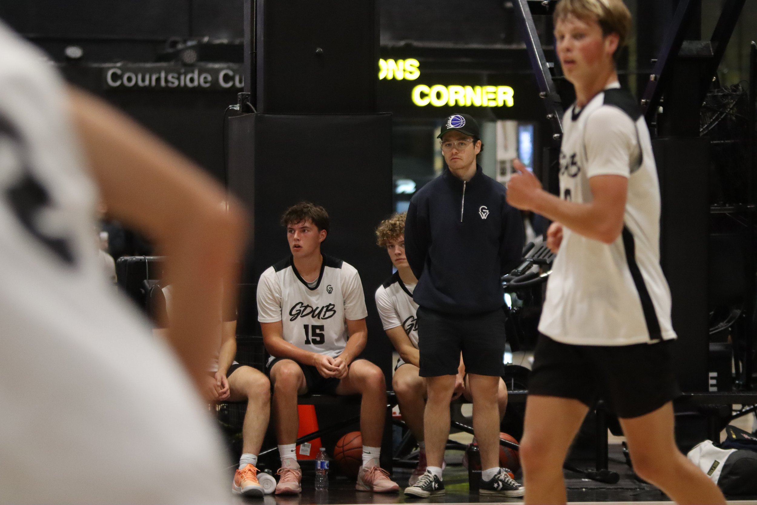 A group of young basketball players sitting and standing on a bench, with a coach or team member speaking to them. The players are wearing white jerseys with black accents and the word 'GQUB' printed on them. One player is wearing a cap, and there are basketballs and water bottles on the ground. The background shows a sign with the word 'CUP' and other text, indicating an indoor sports arena.