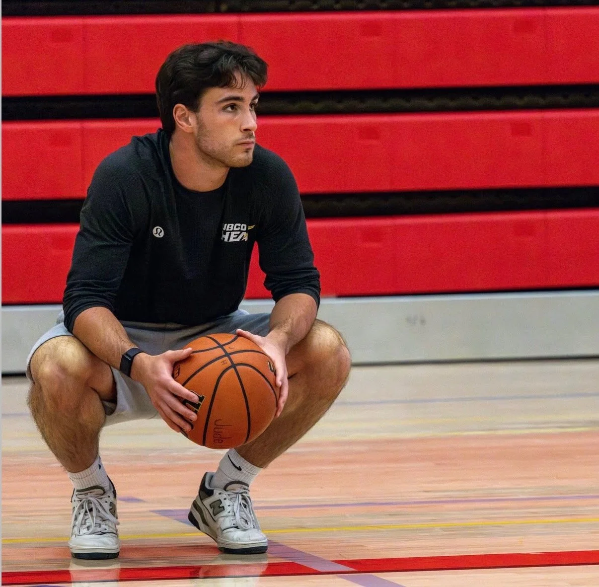 A young man crouching on a basketball court holding a basketball, wearing athletic clothes and sneakers, with red gym bleachers in the background.