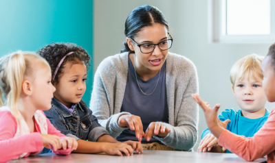 A young teacher wearing glasses and a grey cardigan sitting at a table with four young children, engaged in an active conversation or lesson. One child is gesturing with their hand while the others listen.