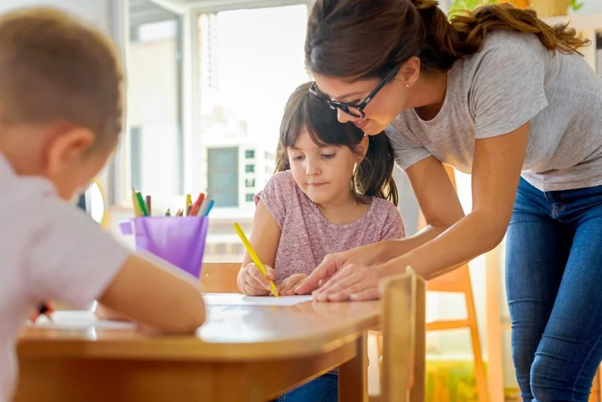 A female teacher with glasses leaning over a wooden desk to help a young girl with her drawing, while another student works in the foreground.