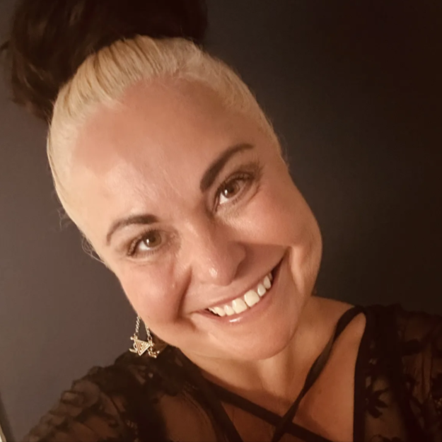 A woman with blonde hair in a bun, smiling and wearing earrings and a black lace top.