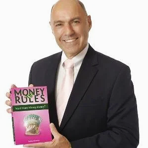 A man in a black suit and pink tie smiling and holding a pink book titled 'Money Rules' with an image of a cupcake on the cover.
