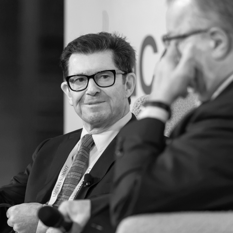 Black and white photo of a dark haired businessman wearing dark glasses in a dark suit on stage