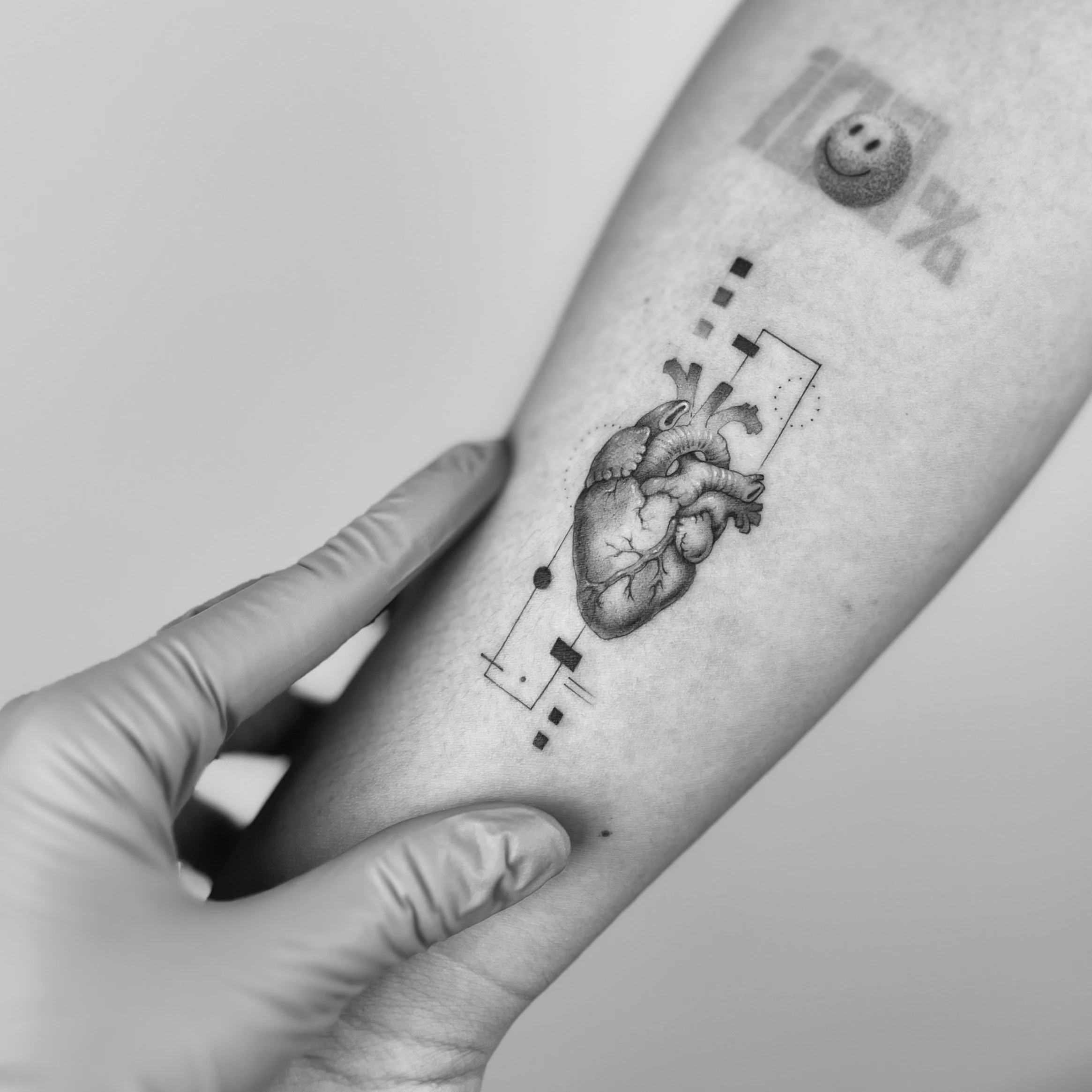 A tattoo of a realistic human heart with geometric and dashed line elements on a person's forearm. A hand wearing a glove touches the tattoo.