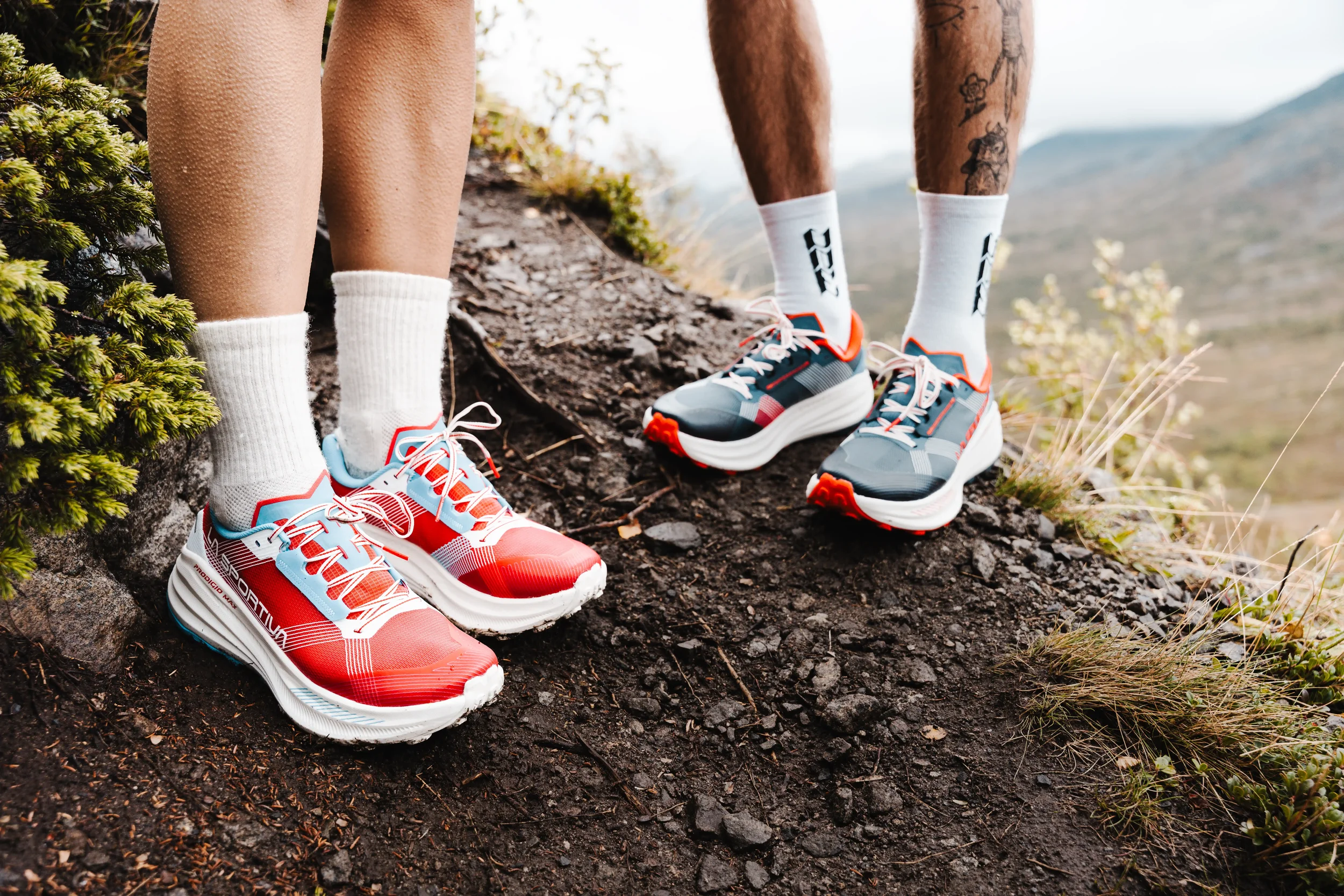 Close-up of two people standing on a rocky trail in athletic shoes and white socks, with a mountain landscape in the background.