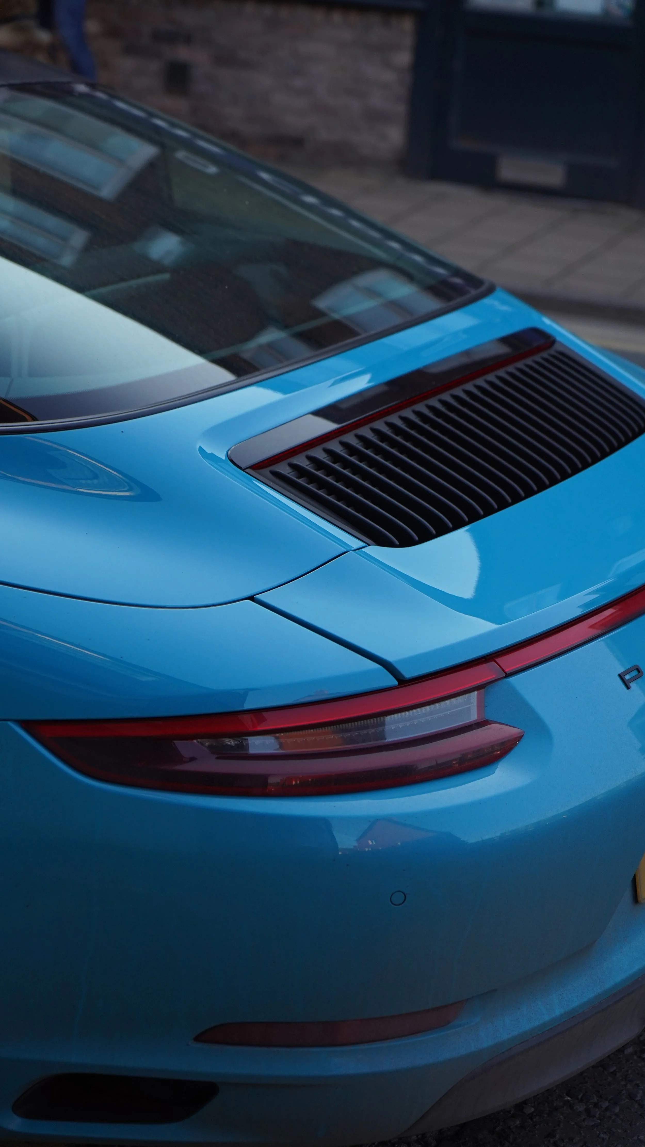 Close-up of the rear end of a blue sports car, showing the tail light, rear windshield, and engine vent.