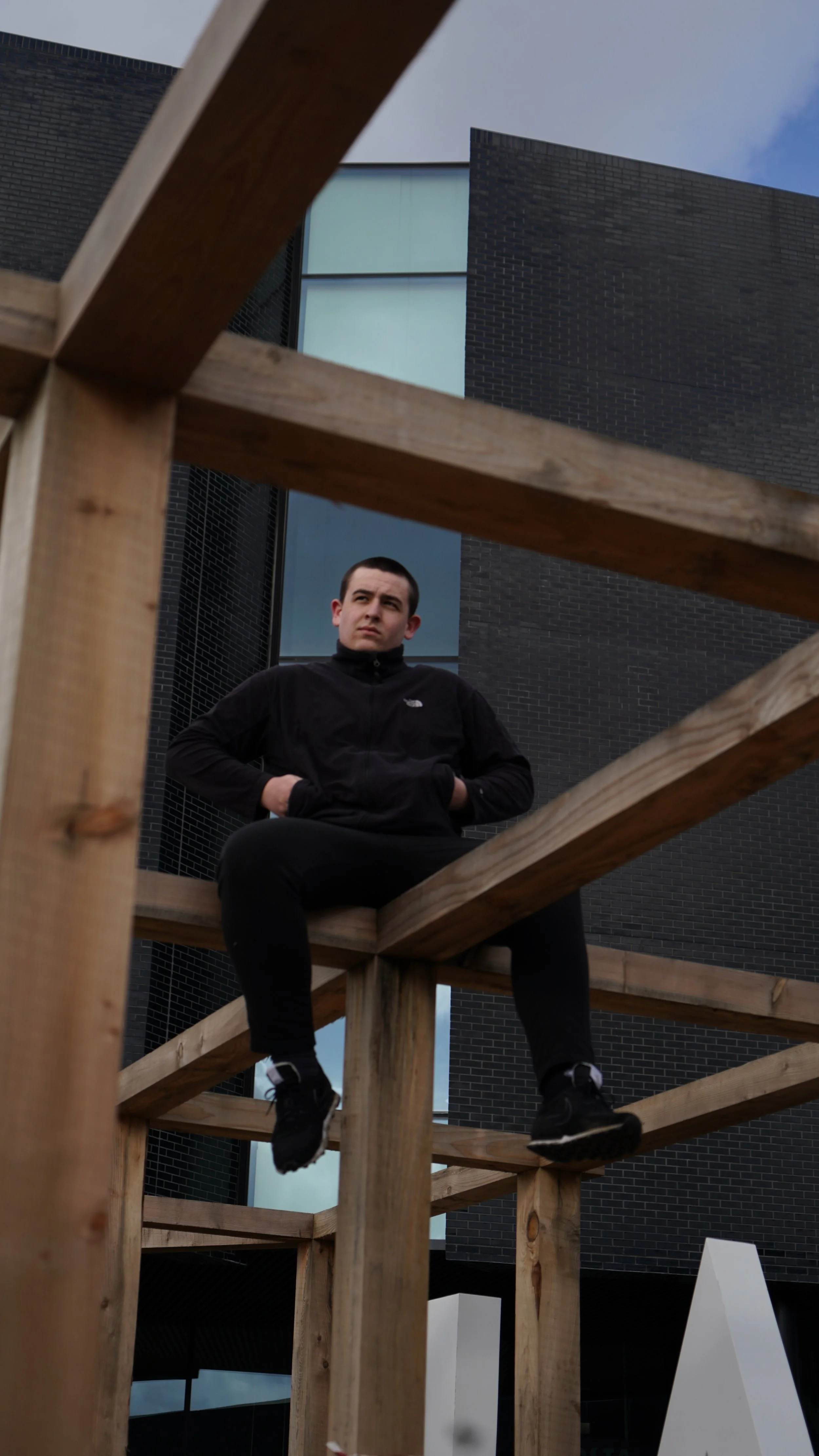 A man sitting on a wooden structure outdoors, with modern black brick buildings and a partly cloudy sky in the background.