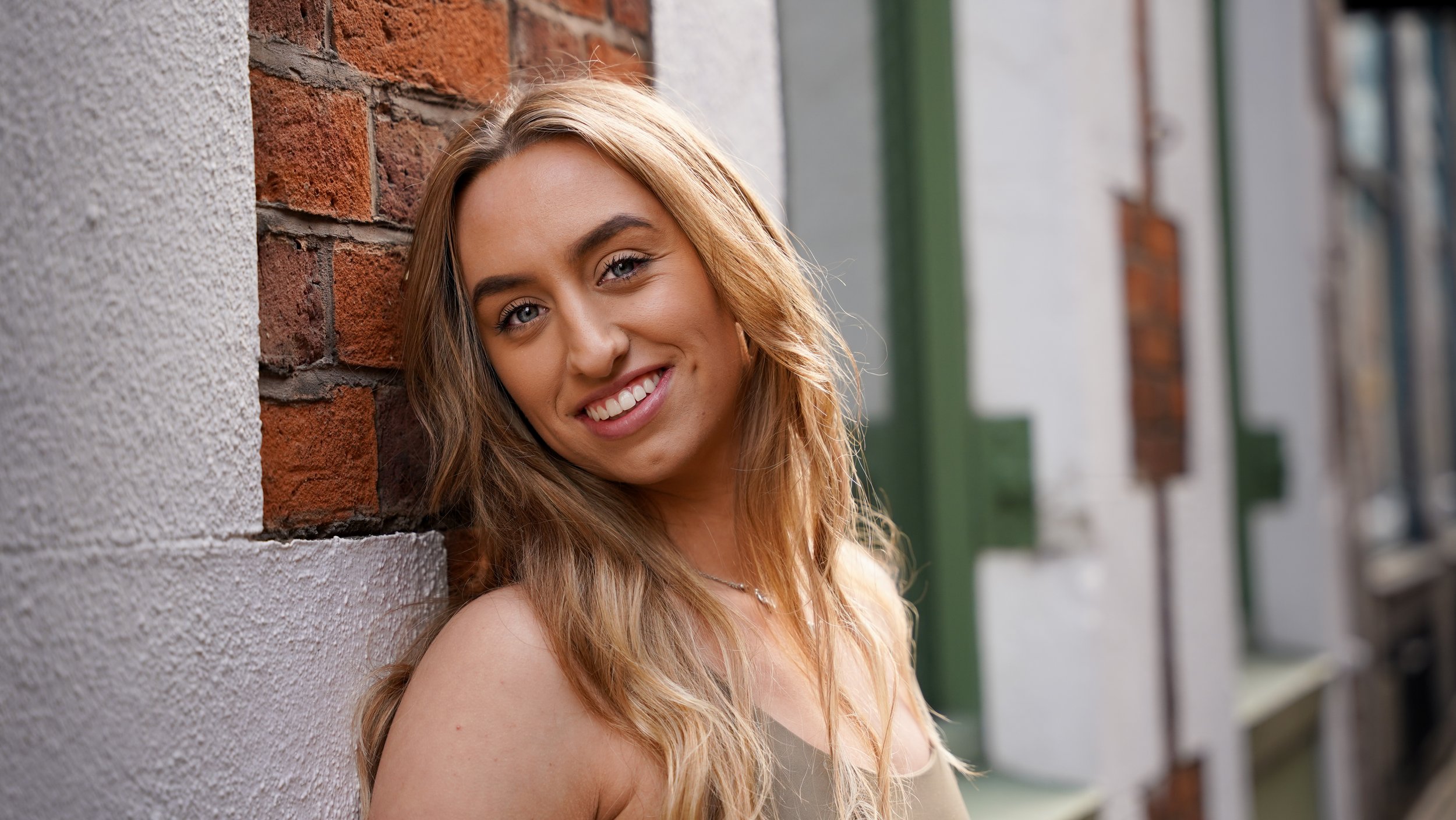 Smiling young woman with long, wavy hair leaning against a brick and concrete wall on a city street.