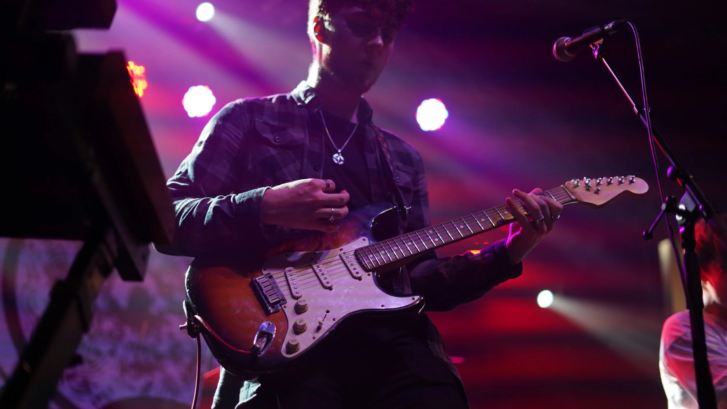 A musician playing an electric guitar on stage with colorful stage lights shining behind him.
