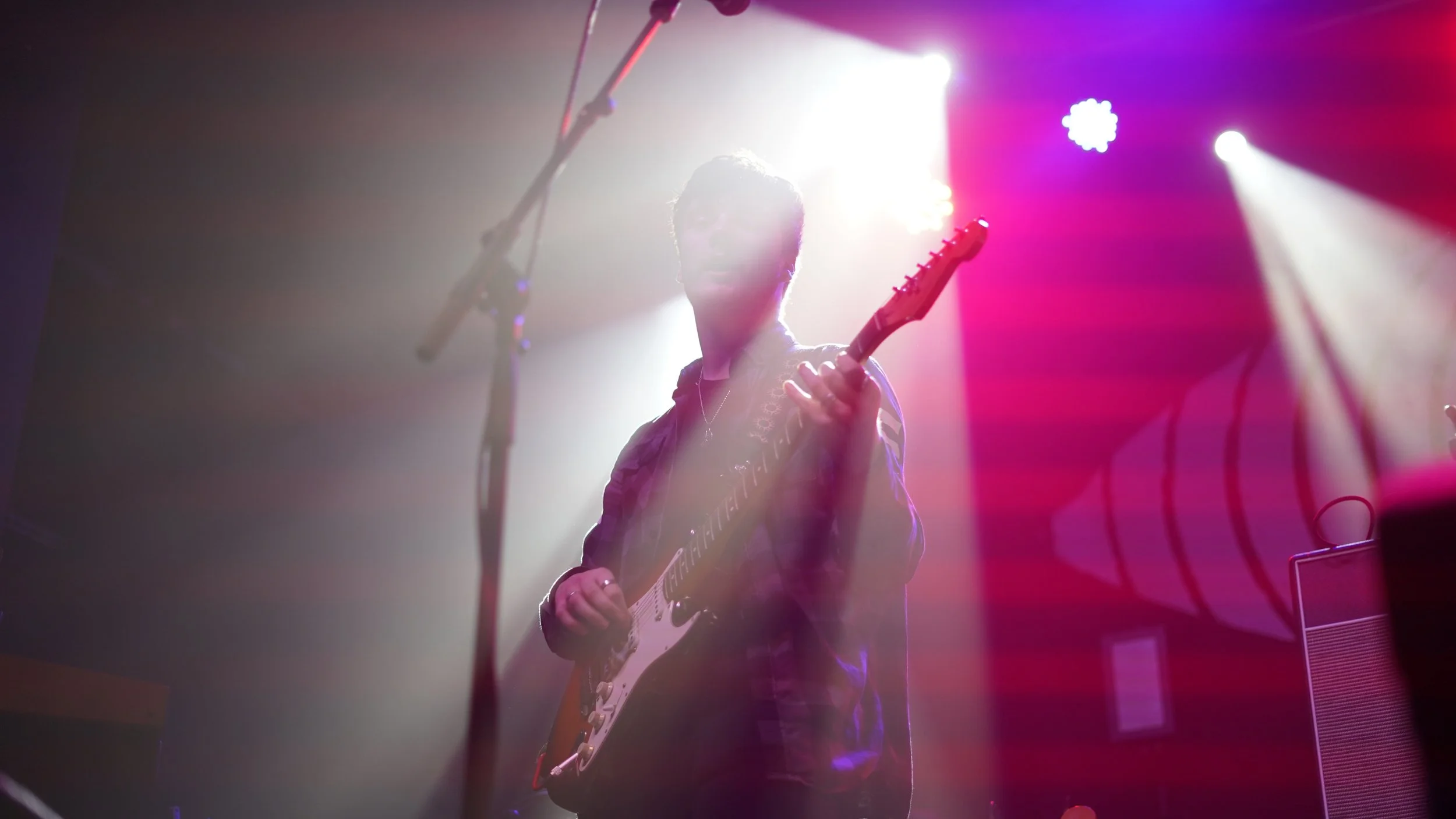 A woman playing an electric guitar on stage with bright backlighting and purple-red stage lights.