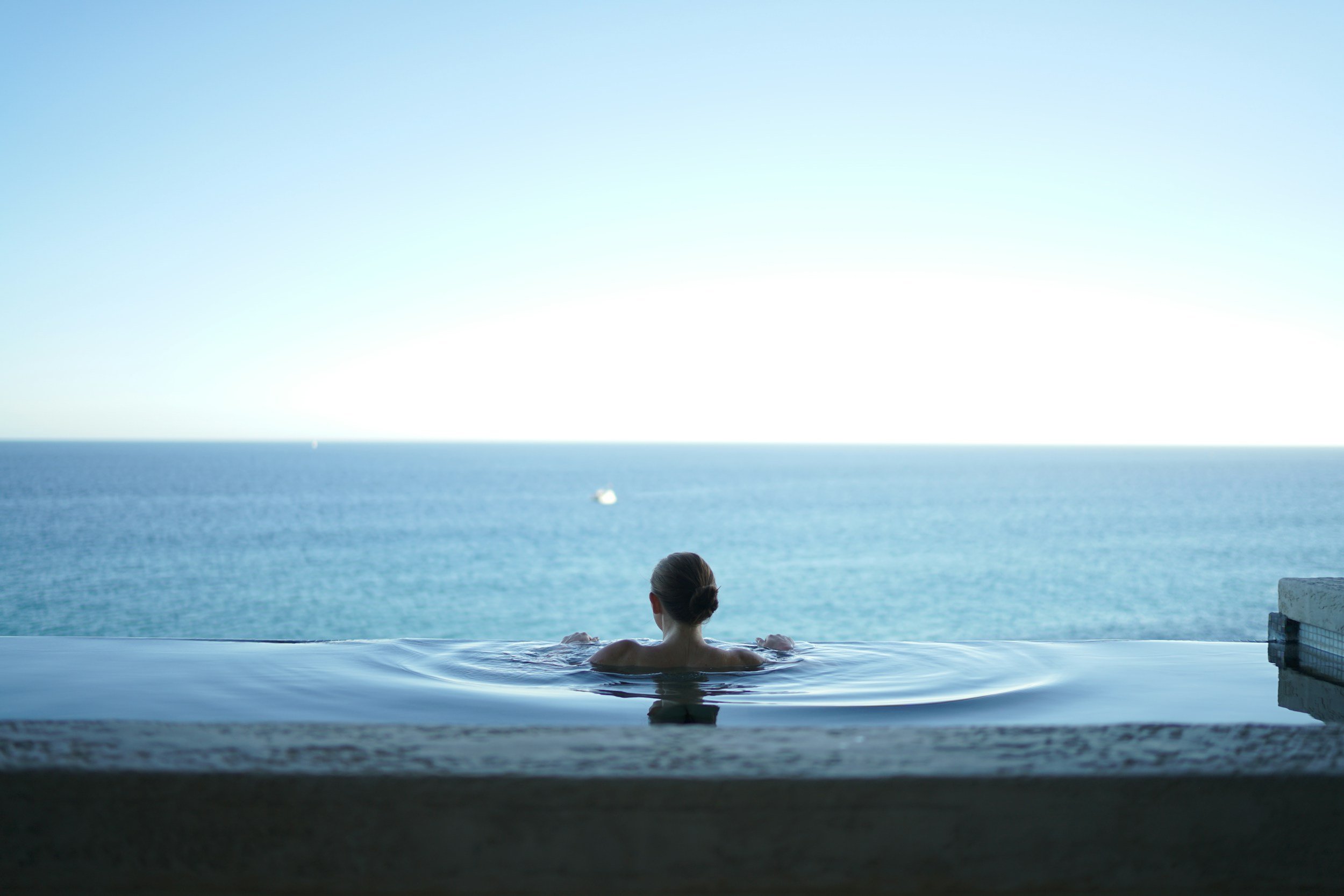Femme nageant dans une piscine à débordement avec vue sur la mer et le ciel bleu.