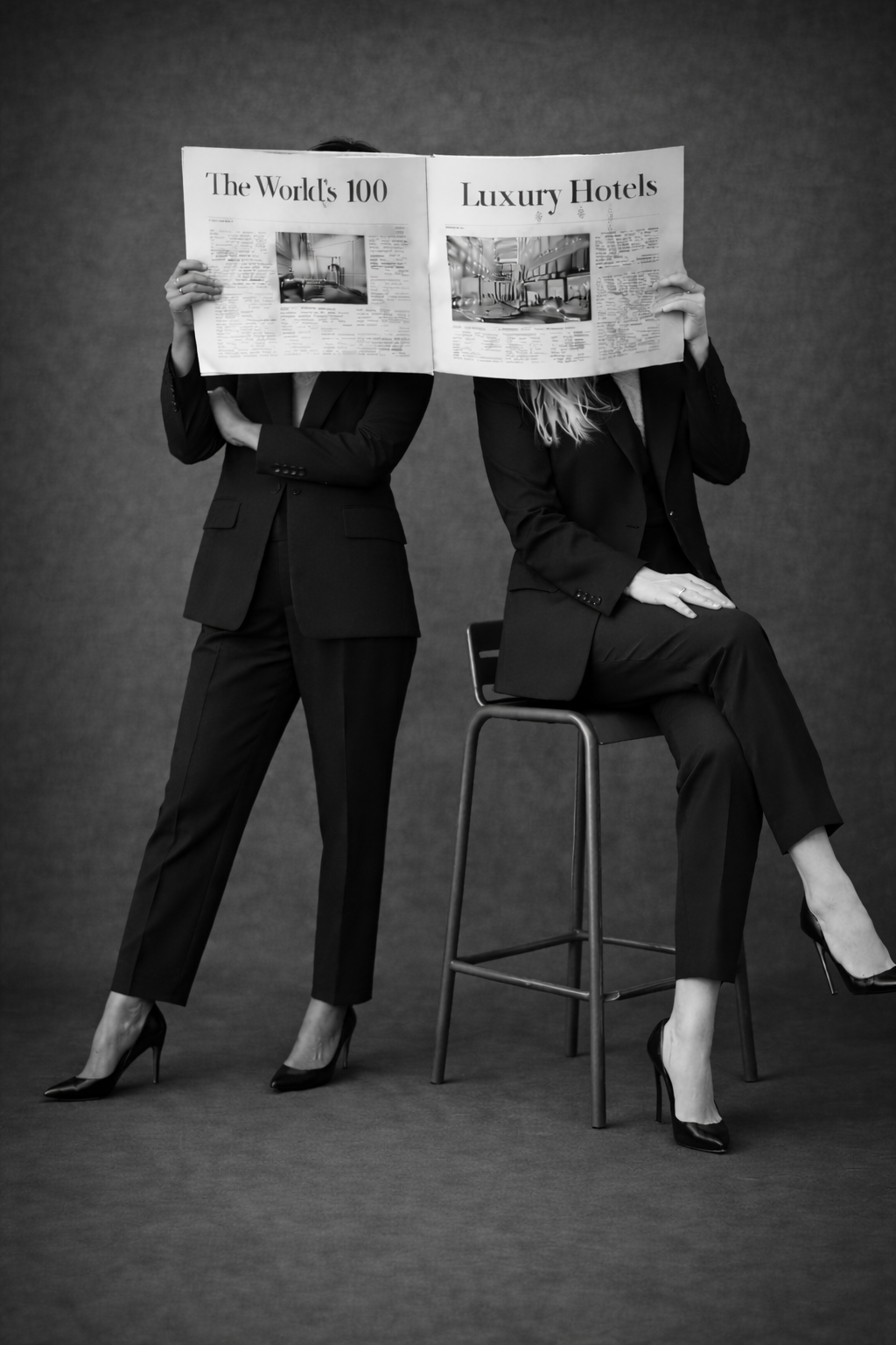 Deux femmes en costume noir, l'une debout et l'autre assise sur une chaise, tenant un journal à leur visage. Le journal a un titre en anglais : 'The World's 100 Luxury Hotels'.