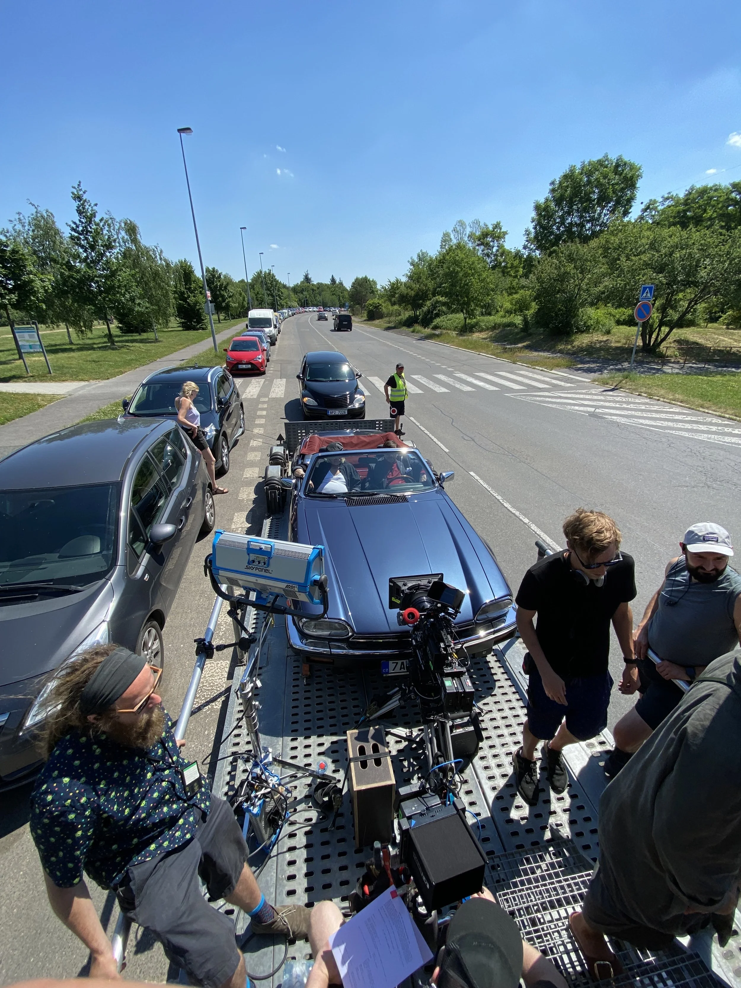 People filming and preparing a scene outdoors with parked cars, trees, and a clear blue sky.